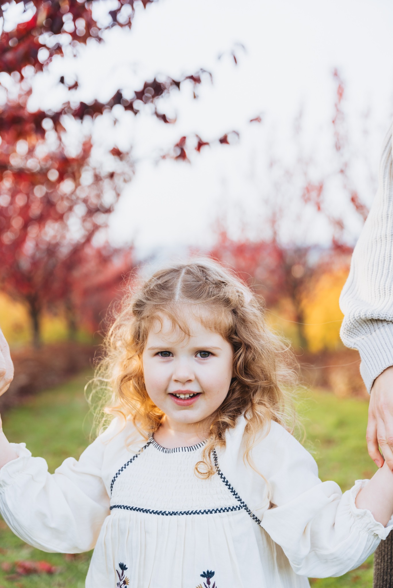 Uśmiechnięta dziewczynka z kręconymi włosami trzymająca za rękę dorosłego na tle drzew z czerwonymi liśćmi w plenerze.