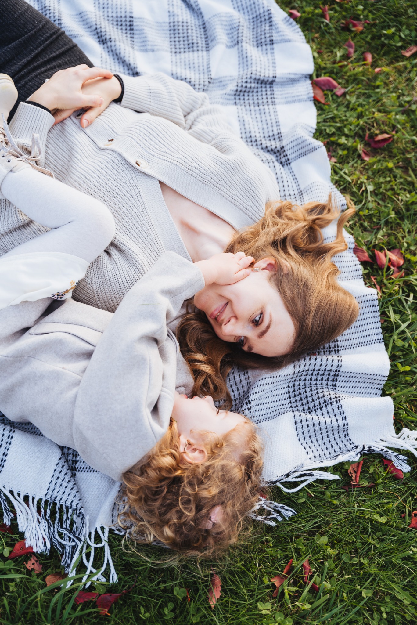 Matka i córka leżą na kocu w kratę na trawie, uśmiechnięte, widok z góry.
