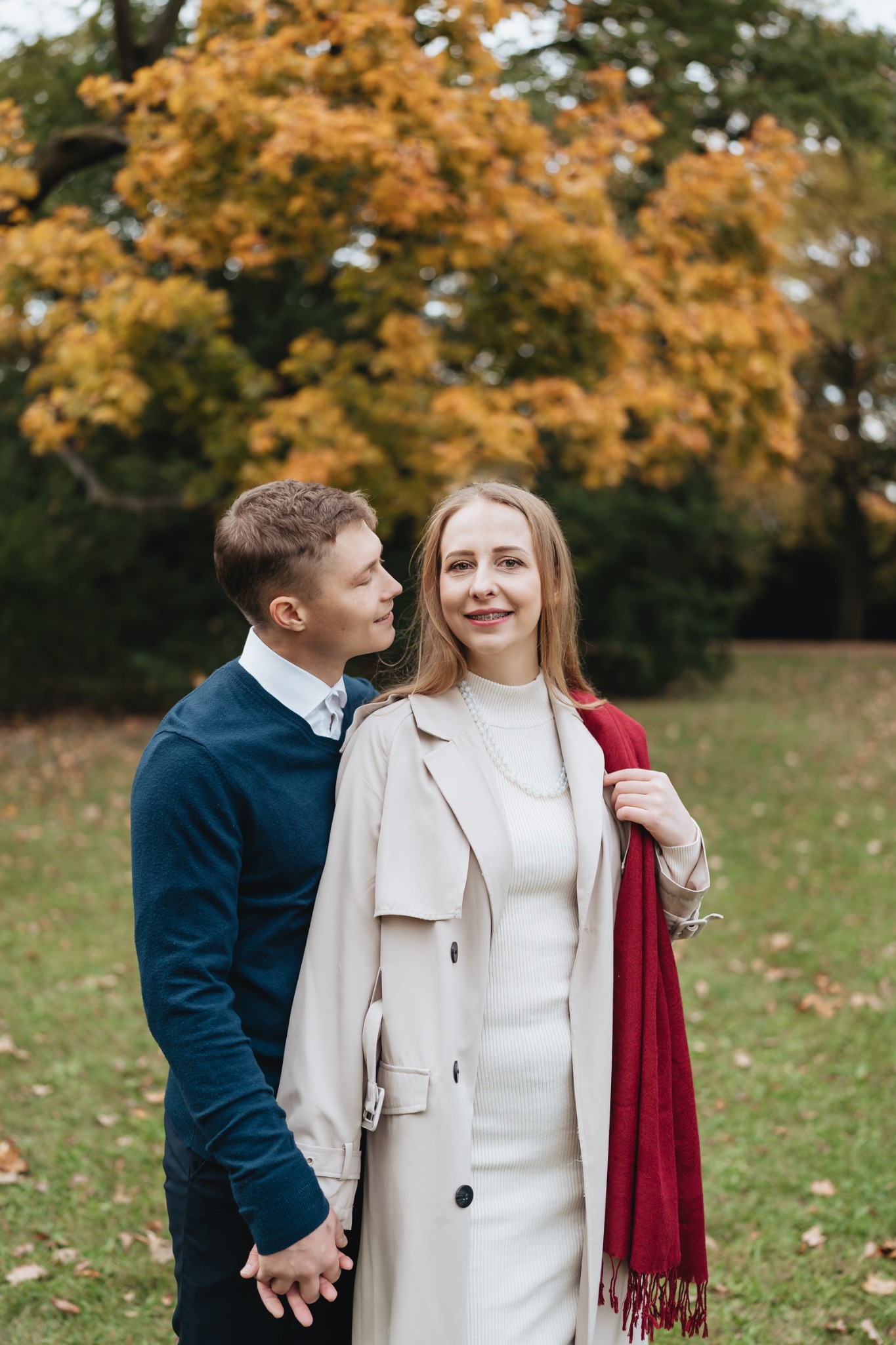 Para trzymająca się za ręce na tle jesiennego parku: mężczyzna w granatowym swetrze spogląda z uśmiechem na kobietę w beżowym płaszczu i białej sukience, z czerwoną chustą na ramieniu i aparatem...