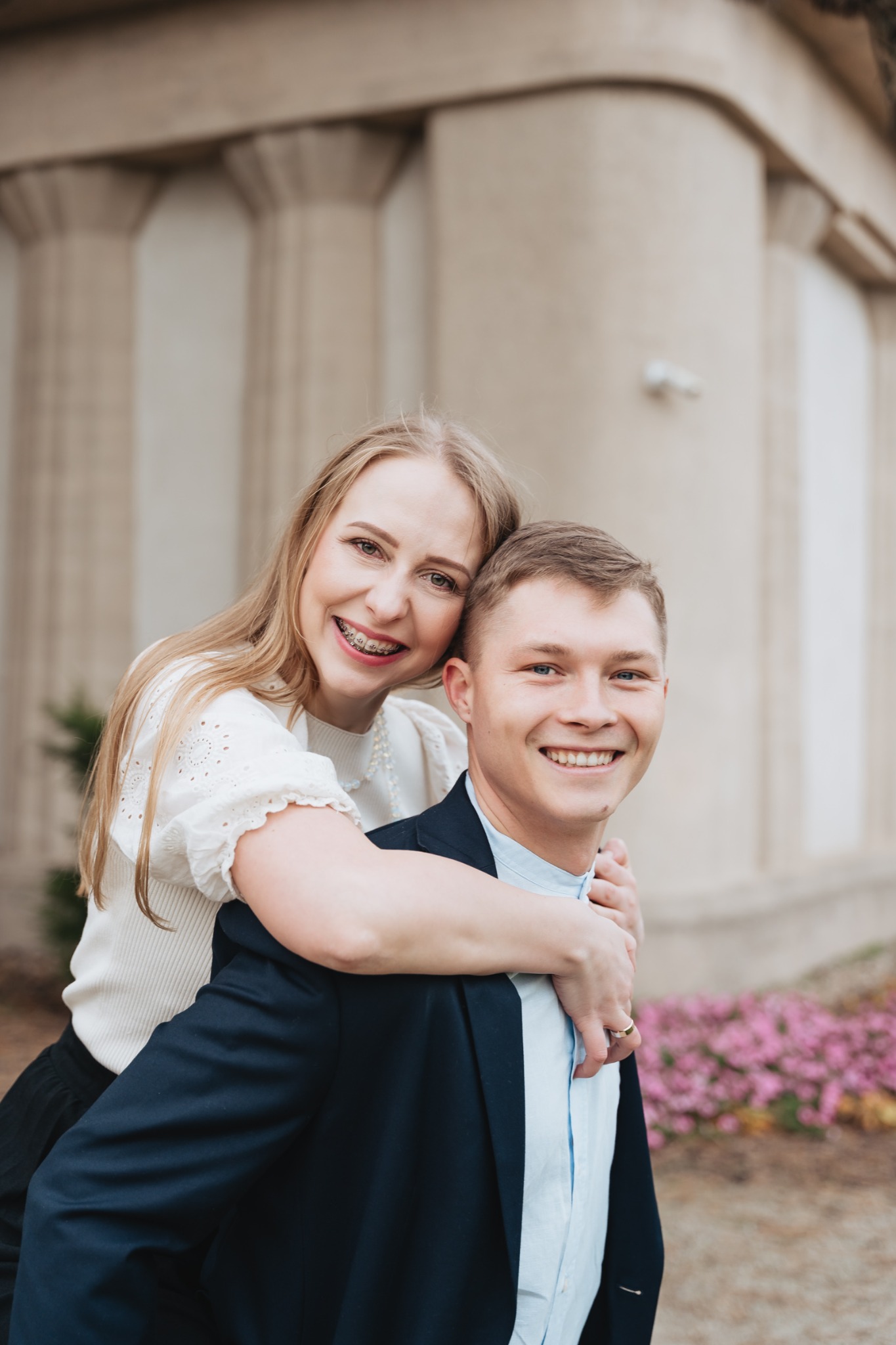 Uśmiechnięta para: blondynka z aparatem ortodontycznym obejmuje mężczyznę w granatowej marynarce na tle budynku z kolumnami i kwitnących różowych kwiatów.