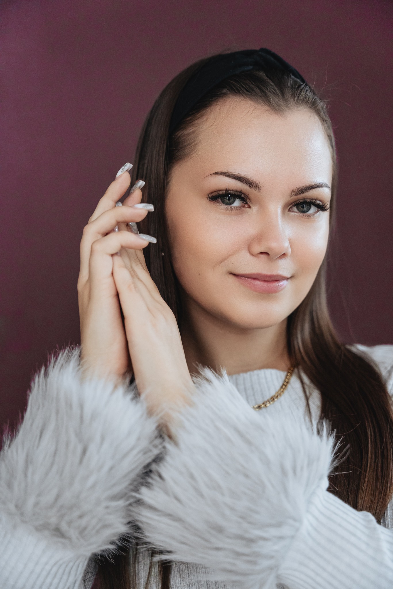 Portret uśmiechniętej brunetki z opaską na włosach, w swetrze z futrzanymi rękawami i złotym łańcuszkiem, dłonie uniesione przy twarzy, paznokcie pomalowane na biało ze srebrnym brokatem.