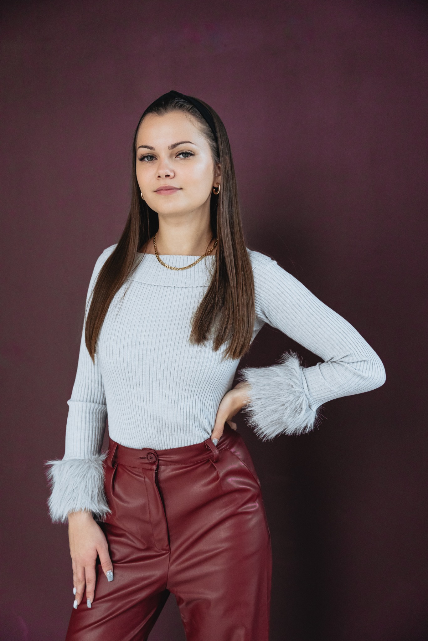 Portret kobiety w prążkowanym swetrze z futrzanymi mankietami i bordowych skórzanych spodniach, na tle śliwkowego tła.