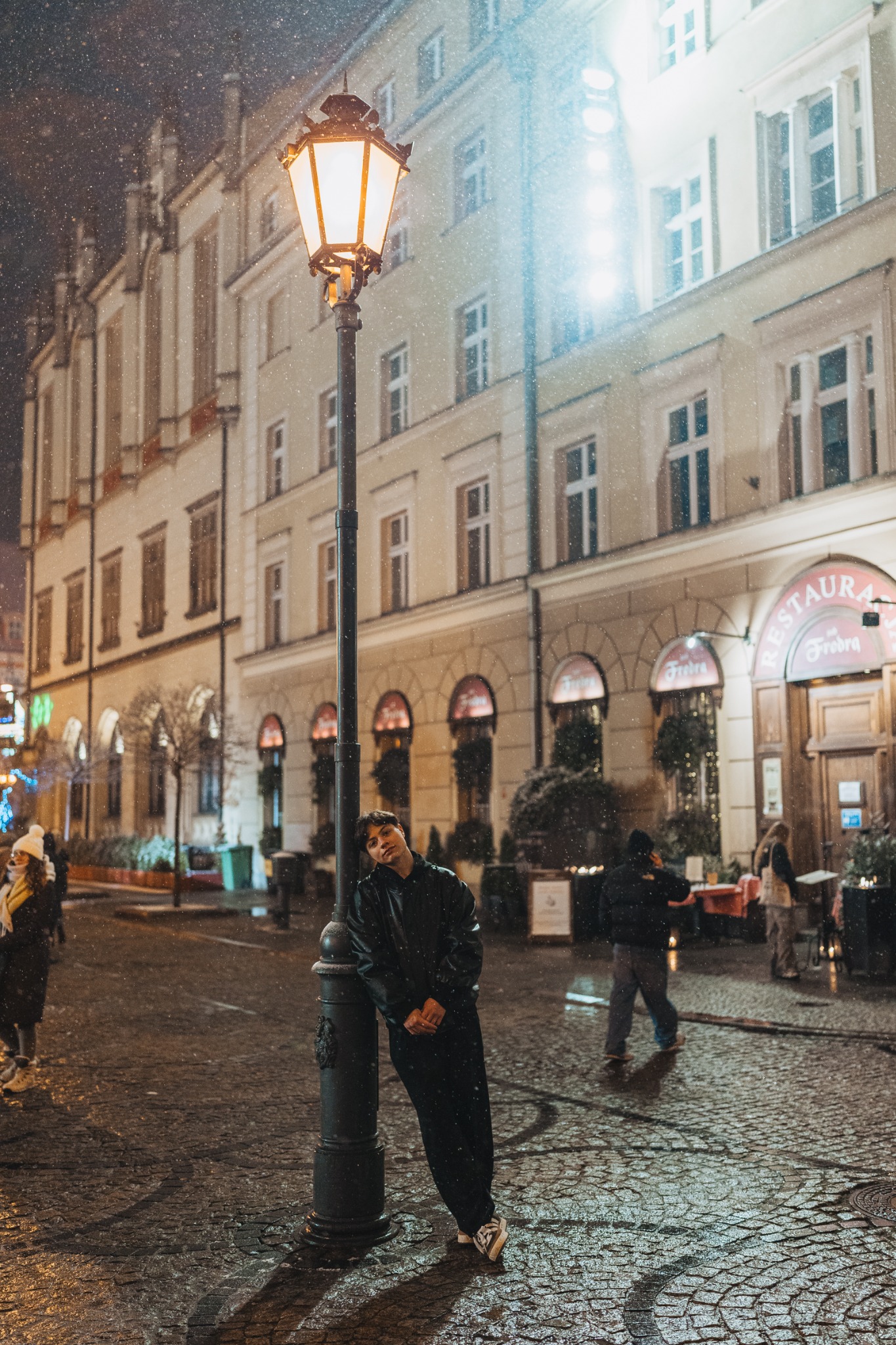 Młody mężczyzna w czarnym ubraniu opiera się o latarnię w śnieżną noc na brukowanej ulicy, w tle oświetlona fasada restauracji.