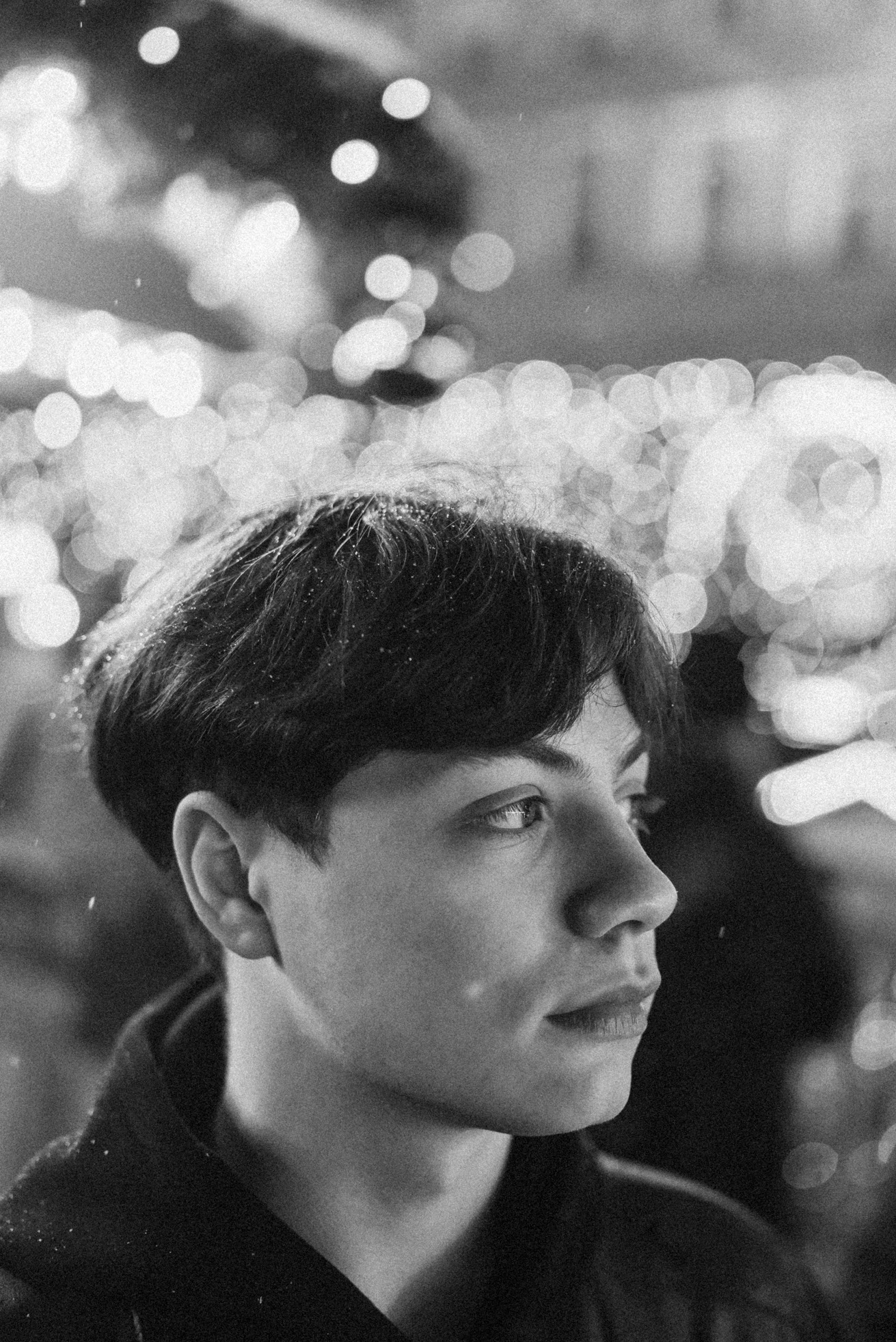 Czarno-biały portret młodego mężczyzny z krótkimi włosami, oświetlonego rozmytymi światłami w tle, z widocznymi drobnymi cząsteczkami na włosach.