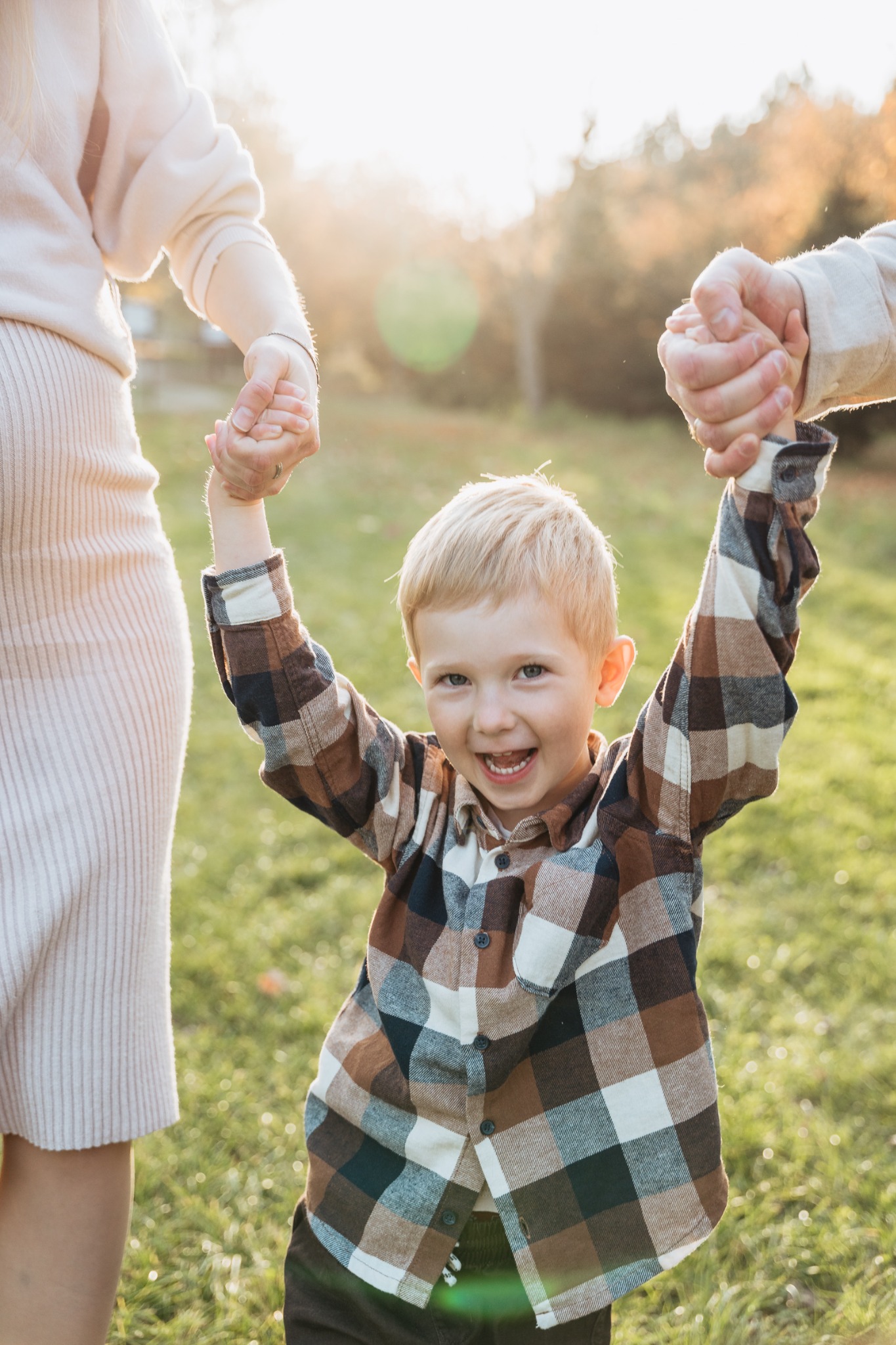 Uśmiechnięty blond chłopiec w brązowo-białej koszuli w kratę trzymany za ręce przez rodziców na tle zielonej trawy i drzew w słoneczny dzień.