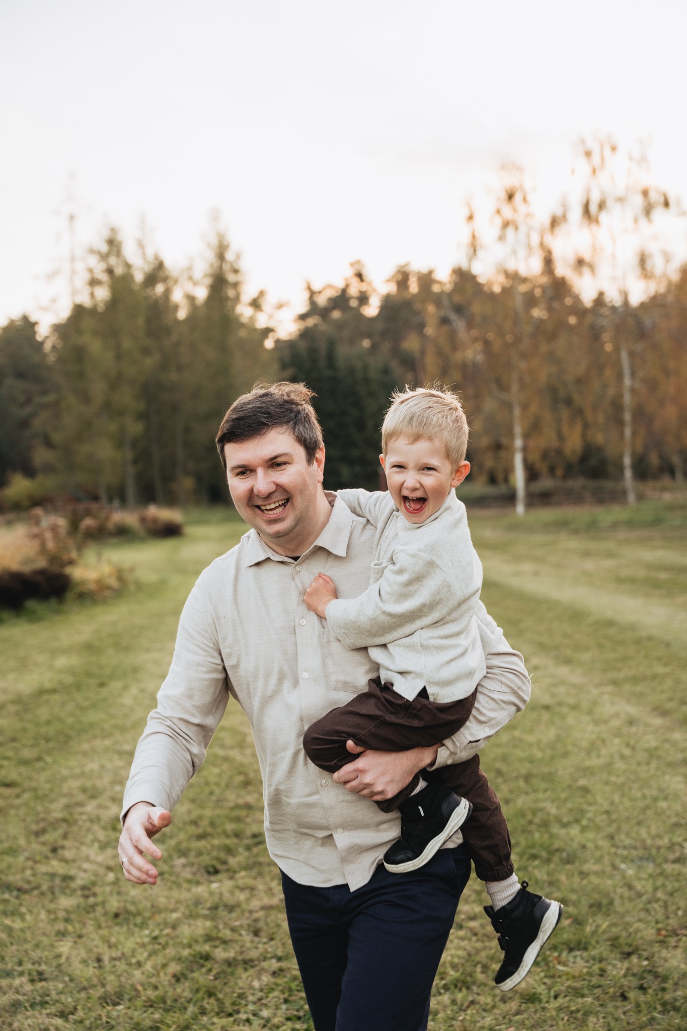 Uśmiechnięty ojciec biegnie po trawie, niosąc na rękach rozbawionego synka w brązowych spodniach i czarnych butach, z otwartą buzią, na tle parku z drzewami.