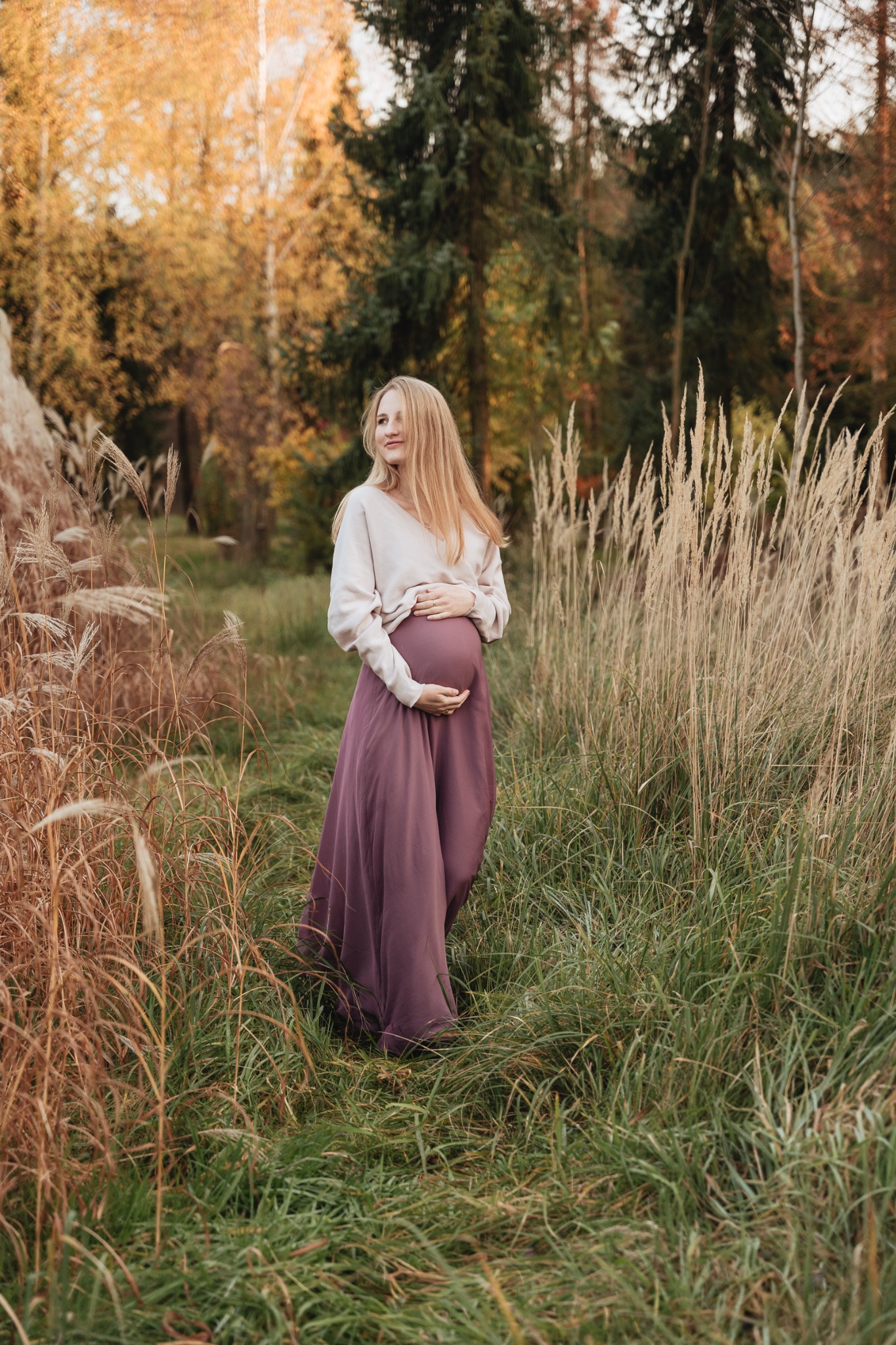 Kobieta w ciąży w długiej fioletowej sukni i jasnym swetrze, pozuje w wysokiej trawie na tle jesiennego lasu, z rękami na brzuchu i delikatnym uśmiechem.