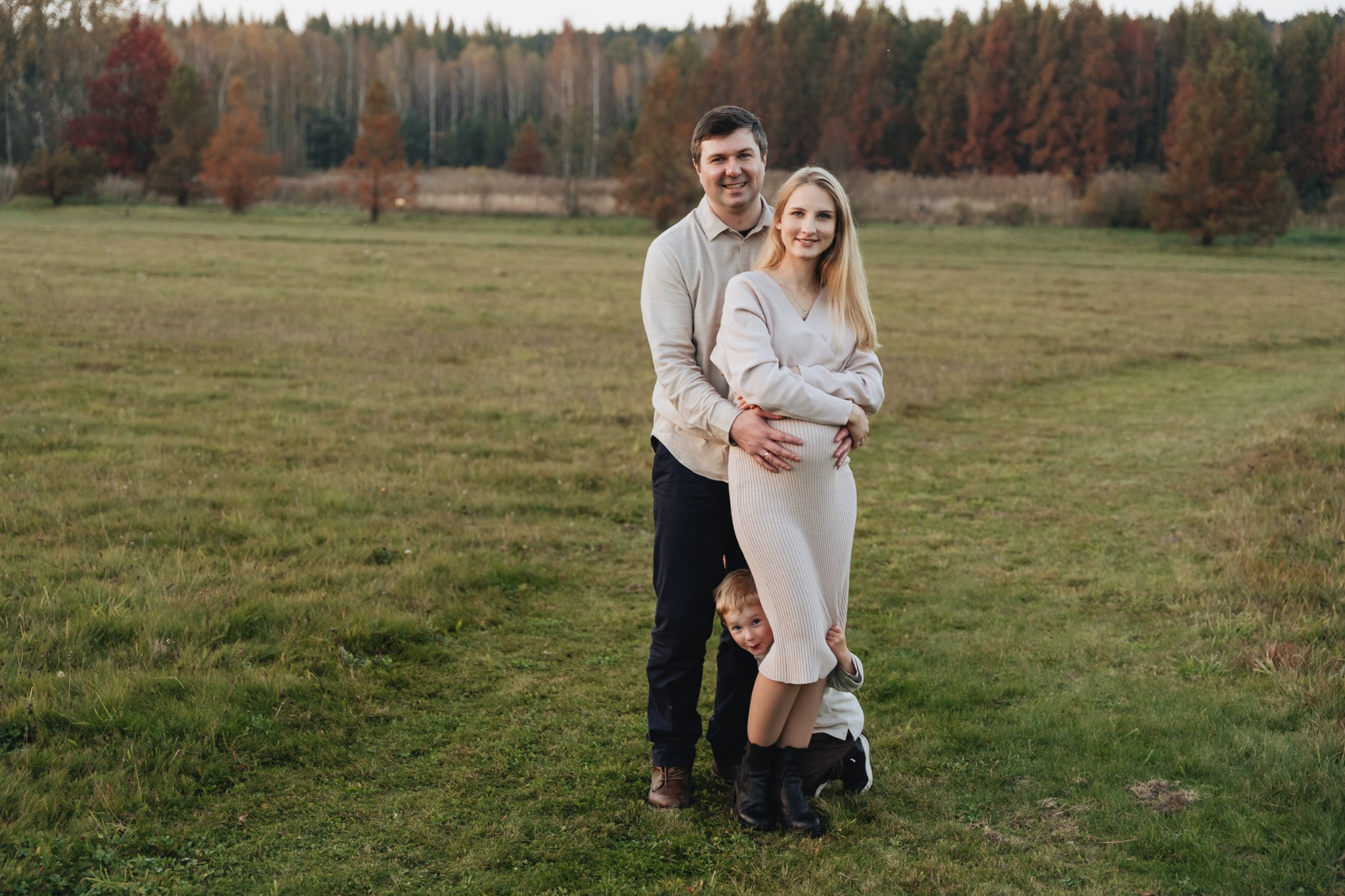 Rodzinne zdjęcie na łące: uśmiechnięty mężczyzna obejmuje ciężarną kobietę w kremowej sukience, zza nóg wystaje mały chłopiec.