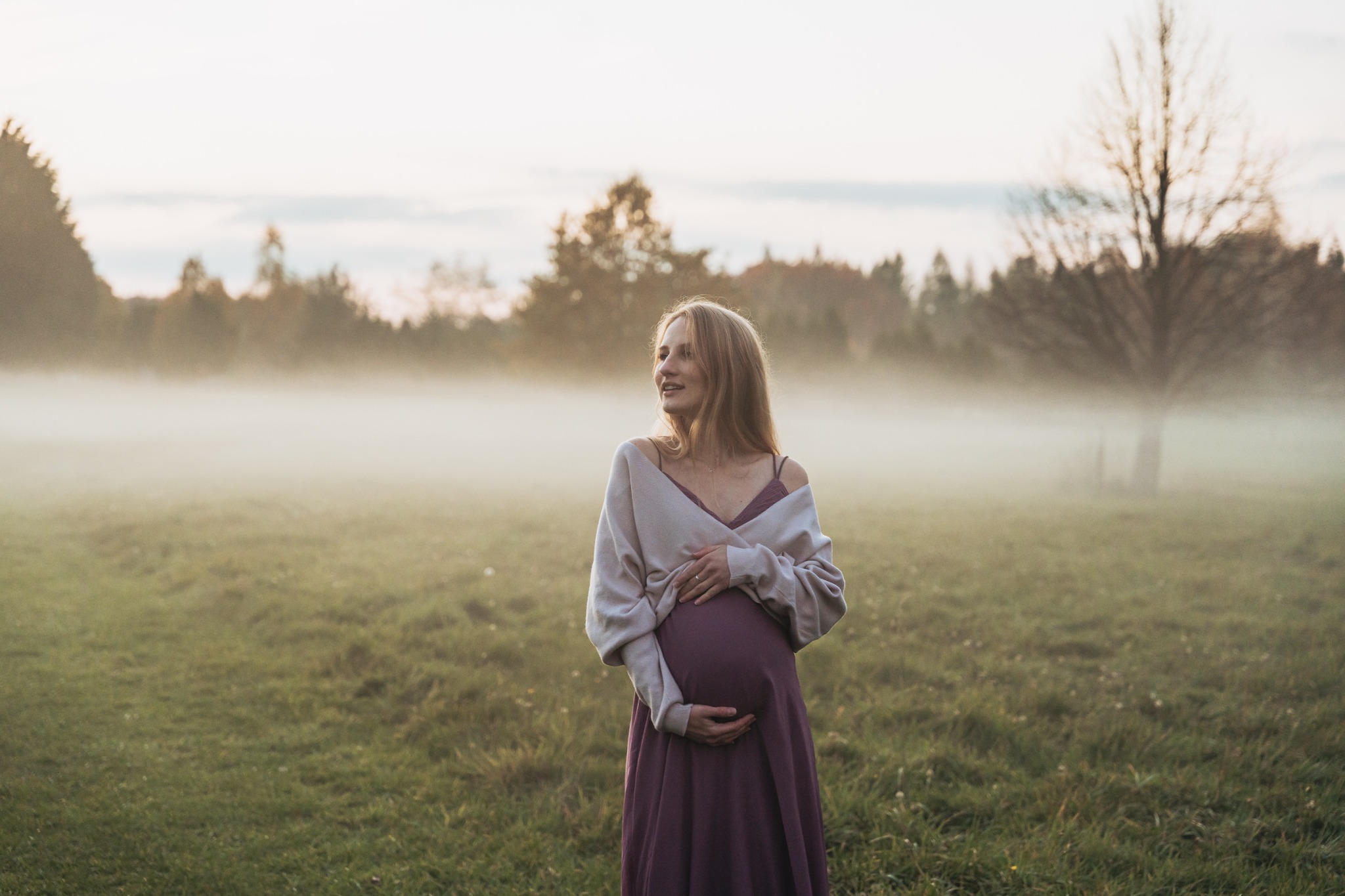 Kobieta w ciąży w długiej sukience i swetrze, pozuje na łące we mgle, z zamyślonym wyrazem twarzy, w tle drzewa.