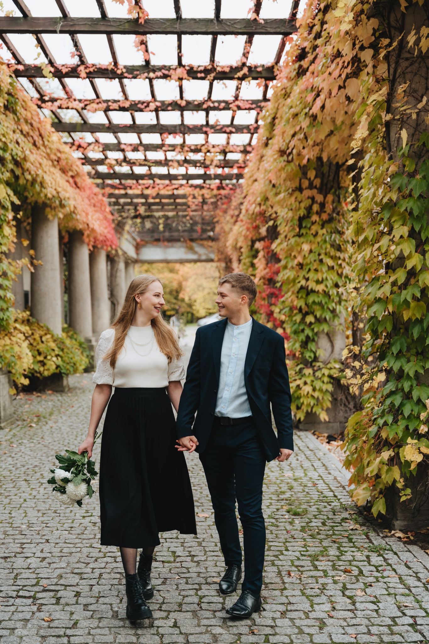 Uśmiechnięta para trzymająca się za ręce, spacerująca po brukowanej ścieżce pod pergolą porośniętą kolorowymi pnączami w jesiennej scenerii. Kobieta w białej bluzce z bufiastymi rękawami, czarnej...