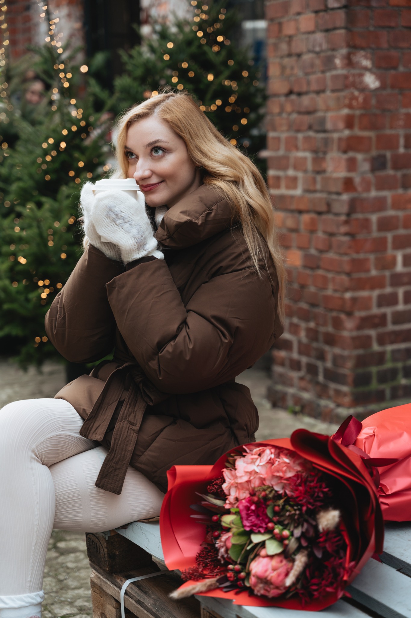 Uśmiechnięta blondynka w brązowej kurtce i białych rękawiczkach pije kawę na wynos na tle ceglanej ściany i choinki z lampkami, obok leży bukiet czerwonych kwiatów.