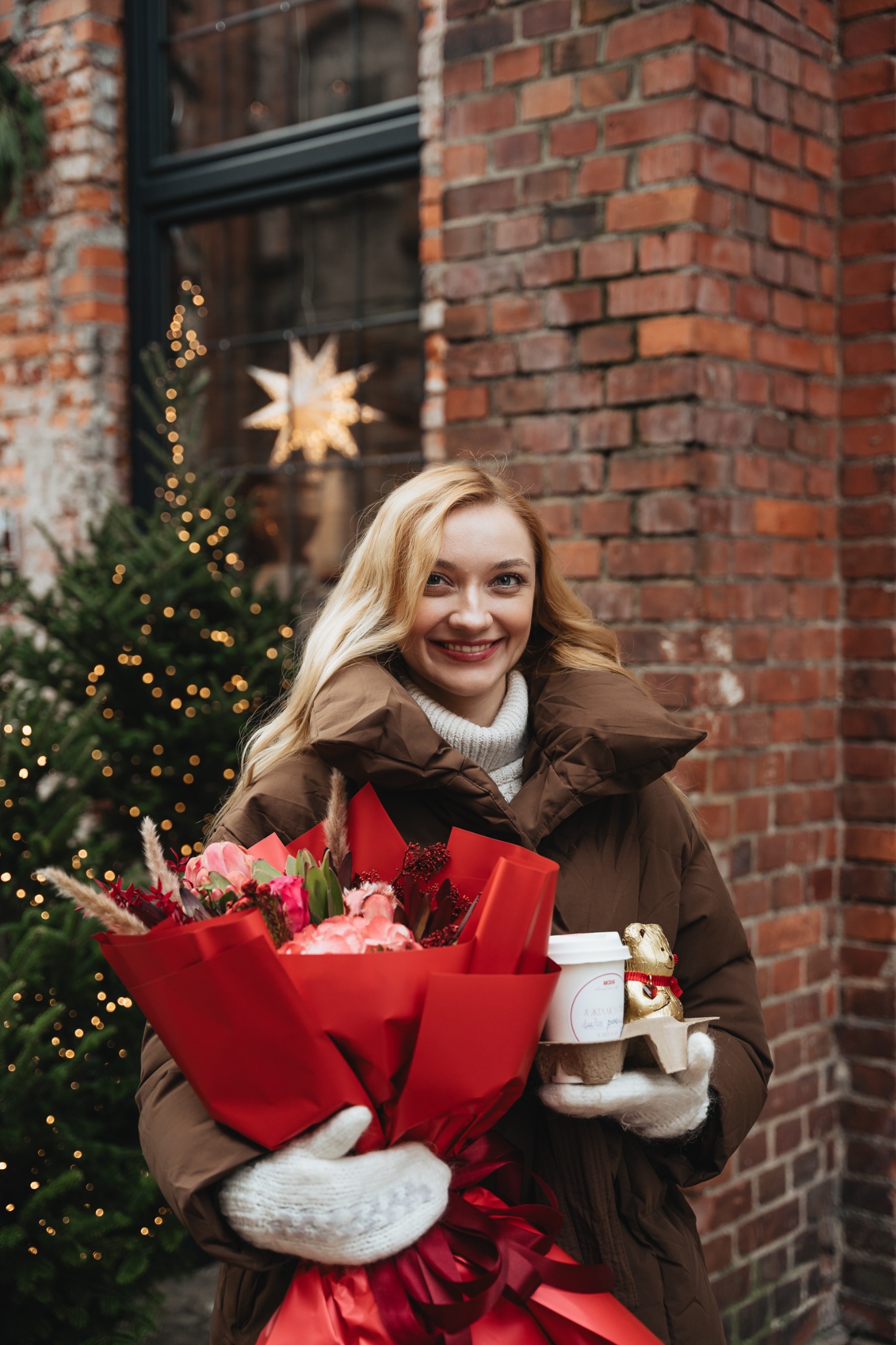 Uśmiechnięta blondynka w brązowej kurtce pozuje z bukietem kwiatów w czerwonym papierze i kawą na tle ceglanej ściany i choinki z lampkami.