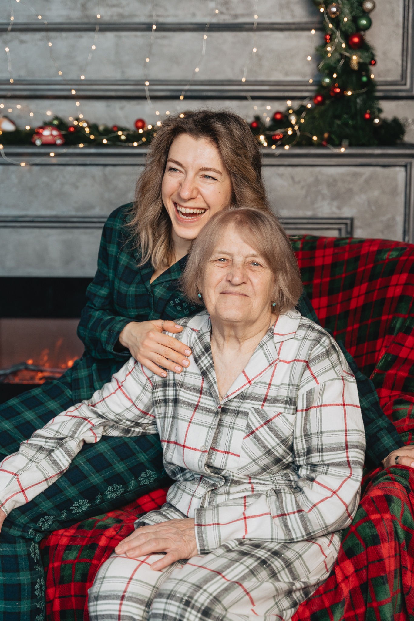 Uśmiechnięta młoda kobieta w zielonej piżamie w kratę obejmuje starszą kobietę w białej piżamie w kratę na tle kominka z dekoracjami świątecznymi.