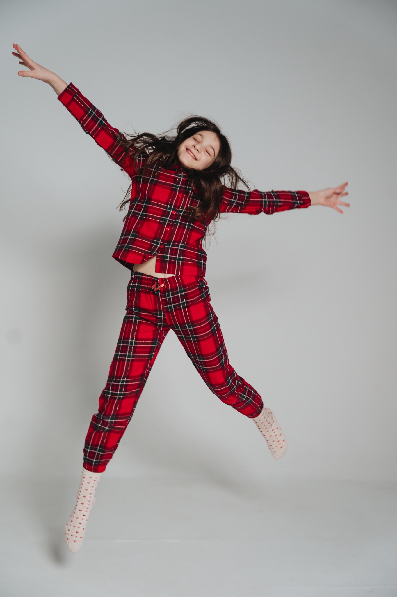 Dziewczynka z zamkniętymi oczami w czerwonej piżamie w kratę skacze na jasnoszarym tle, unosząc ręce w górę.