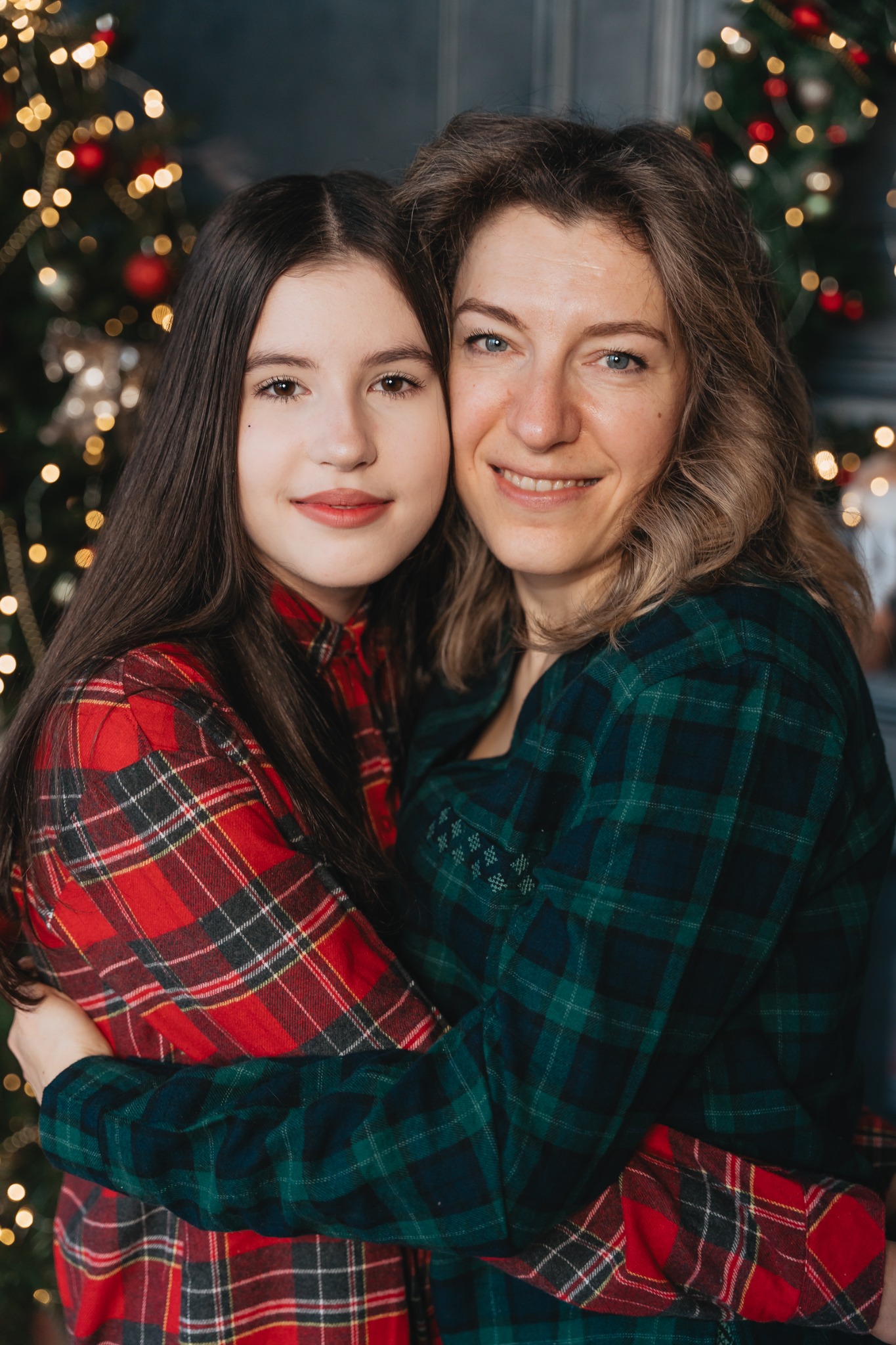 Uśmiechnięta matka i córka w koszulach w kratę obejmują się na tle rozświetlonej choinki.