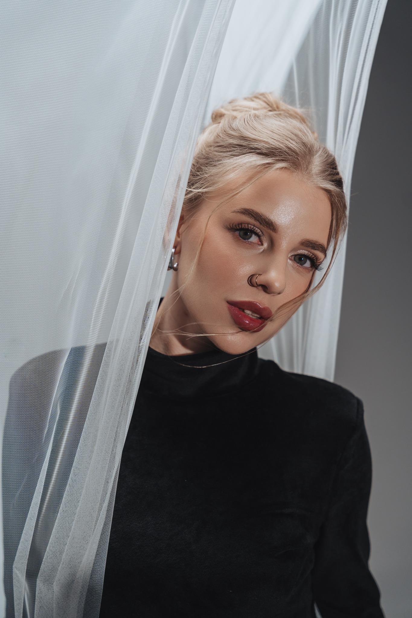 Portret blondwłosej kobiety z piercingiem w nosie, pozującej za delikatną, białą tkaniną na szarym tle.