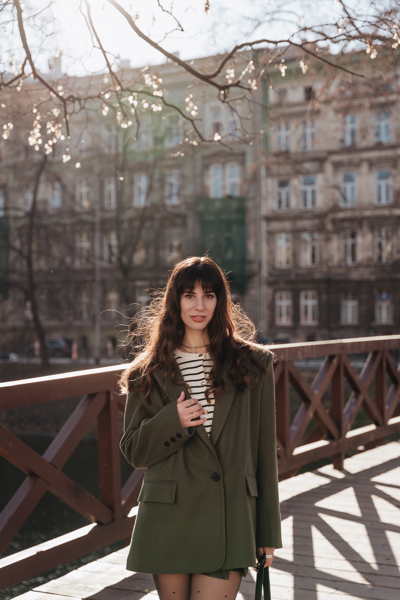 Brunetka w oversize'owym zielonym żakiecie, pozująca na drewnianym moście z miejską zabudową w tle, oświetlona promieniami słońca.