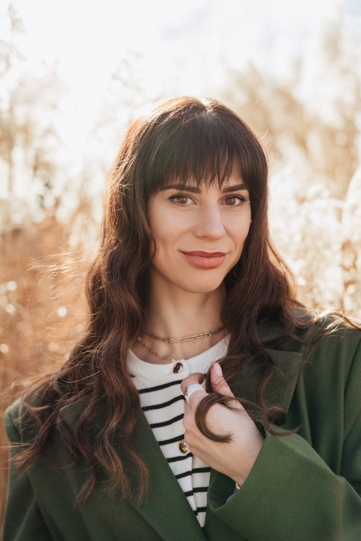 Portret uśmiechniętej kobiety z brązowymi włosami i grzywką, ubrana w zielony płaszcz i sweter w paski na tle rozmytej roślinności w ciepłych barwach.