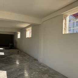 ANT-POL Jakub Glebkowicz - Wnętrze po tynkowaniu: jasne ściany, betonowa podłoga z refleksami światła słonecznego wpadającego przez okna z widokiem na budynek z czerwonym dachem.