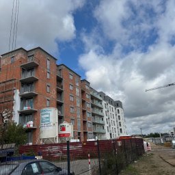 ANT-POL Jakub Glebkowicz - Budowa bloku mieszkalnego z cegły, widoczne rusztowania, zbiorniki na tynk i dźwig. Częściowo ocieplony budynek z balkonami na tle błękitnego nieba.