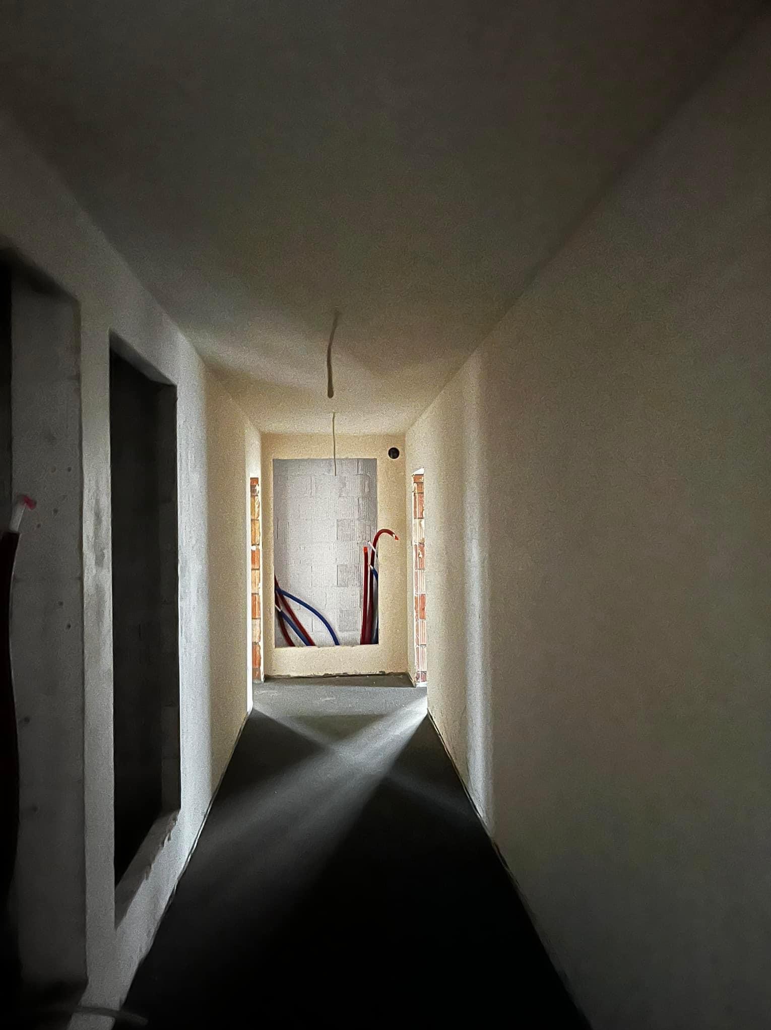 Korytarz w trakcie remontu z surowymi tynkami na ścianach, widoczne instalacje w otworze drzwiowym, ciemna podłoga wylewka.