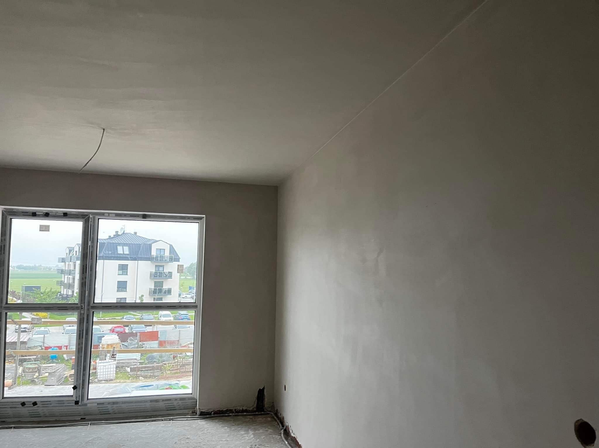 Wnętrze pokoju po tynkowaniu maszynowym, widoczne gładkie ściany i sufit, okno z widokiem na budynek mieszkalny i plac budowy na zewnątrz.