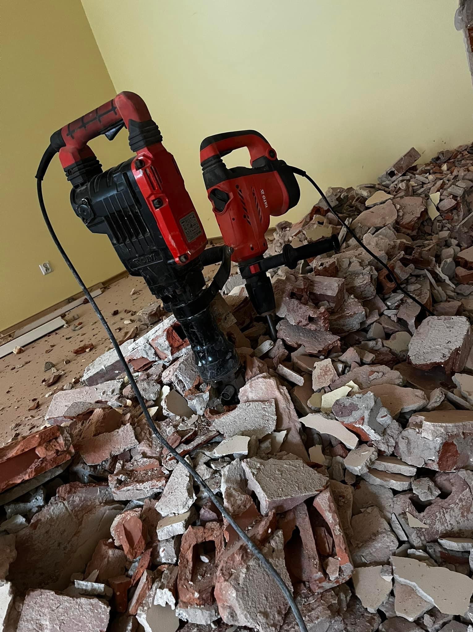 Dwa młoty udarowe Yato na tle gruzu po skuciu tynku ze ściany w pomieszczeniu z żółtymi ścianami i widocznym gniazdkiem elektrycznym.