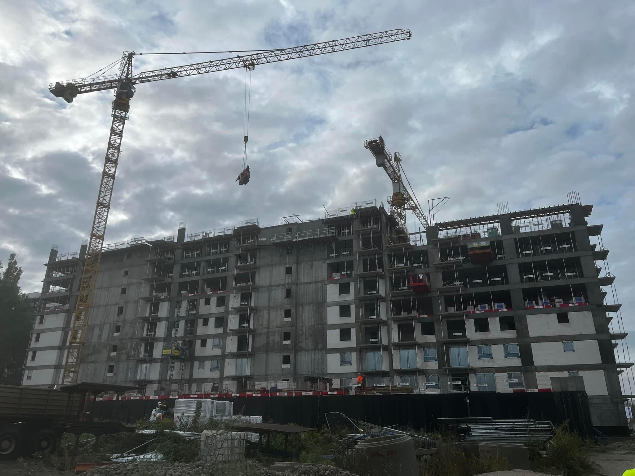 Wielopiętrowy budynek w stanie surowym zamkniętym z dwoma żurawiami budowlanymi na tle pochmurnego nieba, widoczne elementy rusztowań i materiały budowlane na placu budowy.