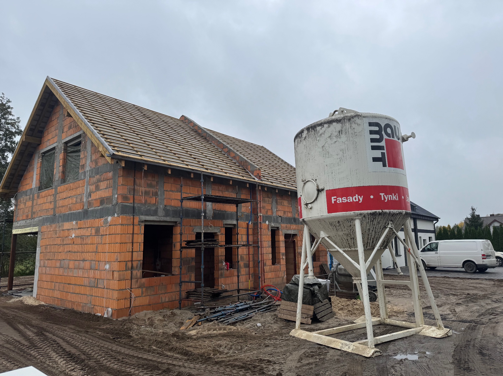 Budowa domu jednorodzinnego w stanie surowym otwartym z widocznym zbiornikiem na tynk maszynowy. Na dachu ułożona dachówka. Widoczne rusztowania.