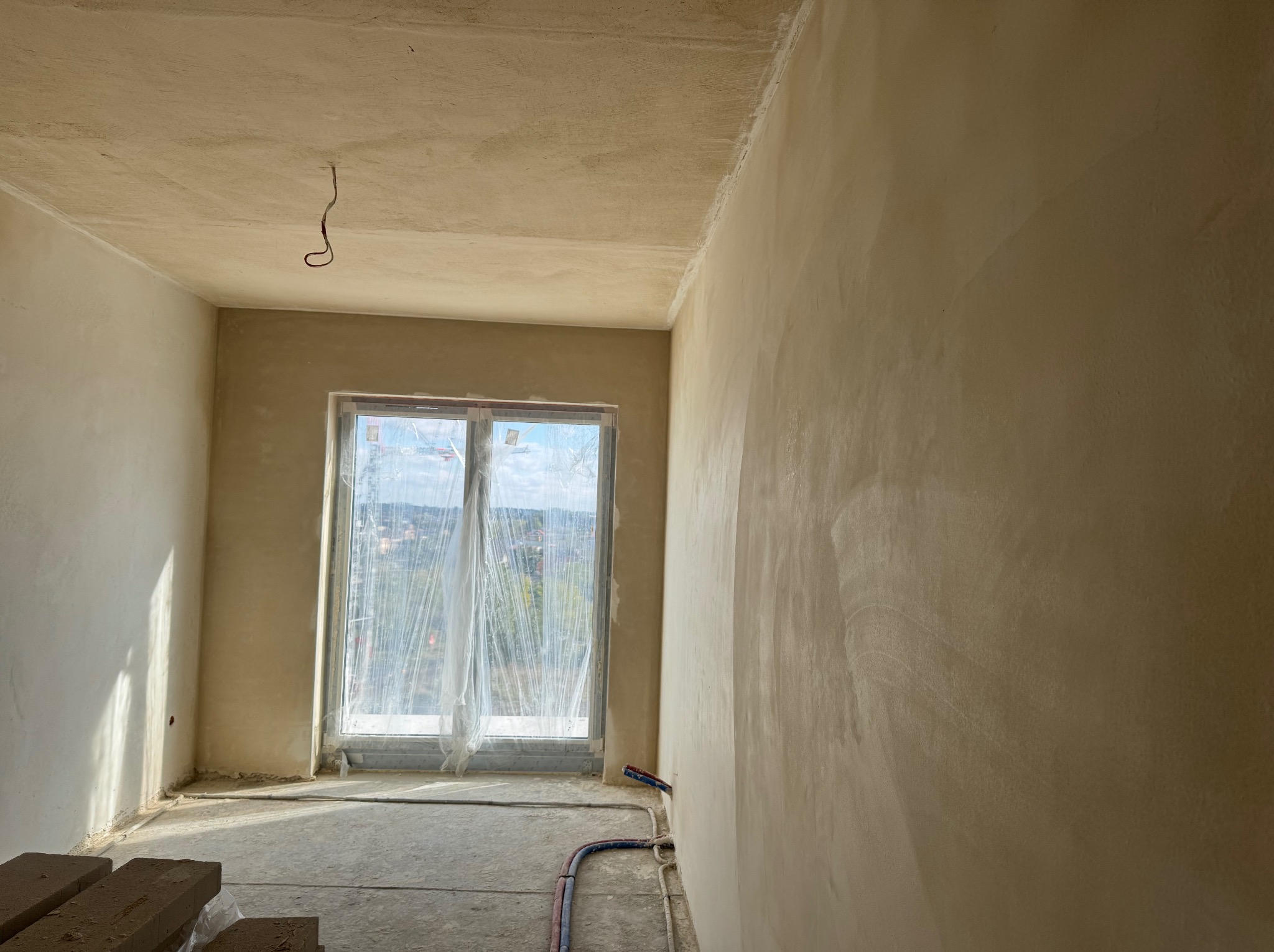 Pokój po tynkowaniu maszynowym: gładkie ściany w odcieniach beżu, okno z widokiem na zewnątrz, przewody na podłodze i cegły w rogu. Widoczne ślady po tynkowaniu.