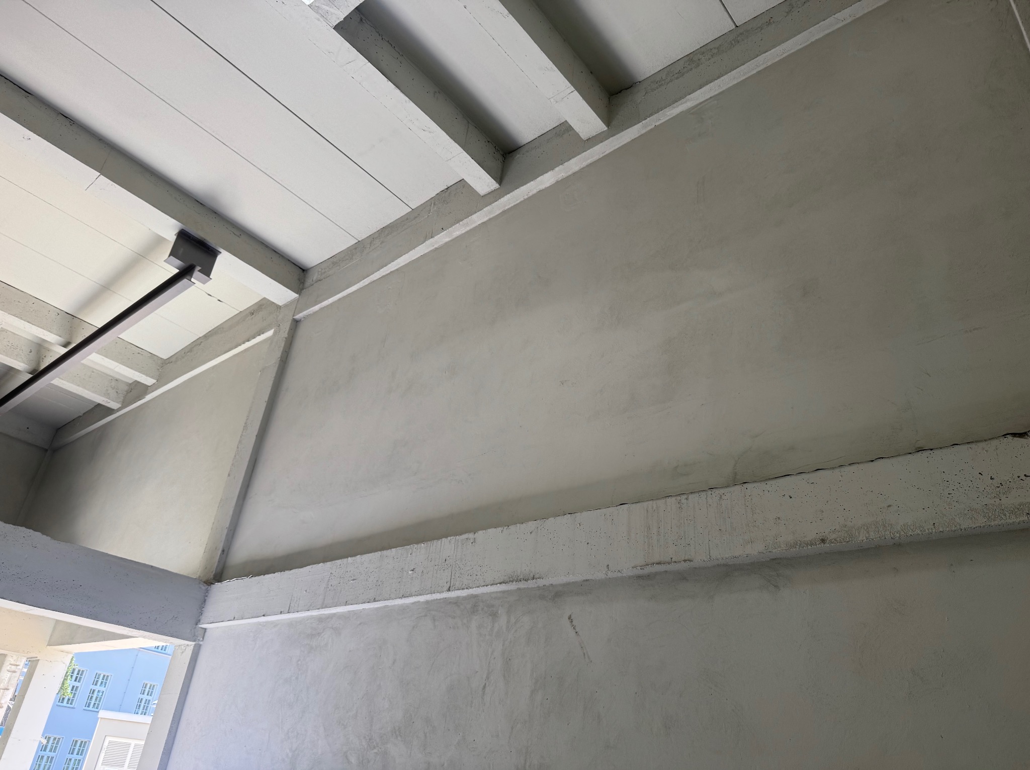 Ściana po tynkowaniu maszynowym z widocznymi elementami konstrukcyjnymi stropu i słupów. Szara, gładka powierzchnia, surowy beton. Widok z dołu.