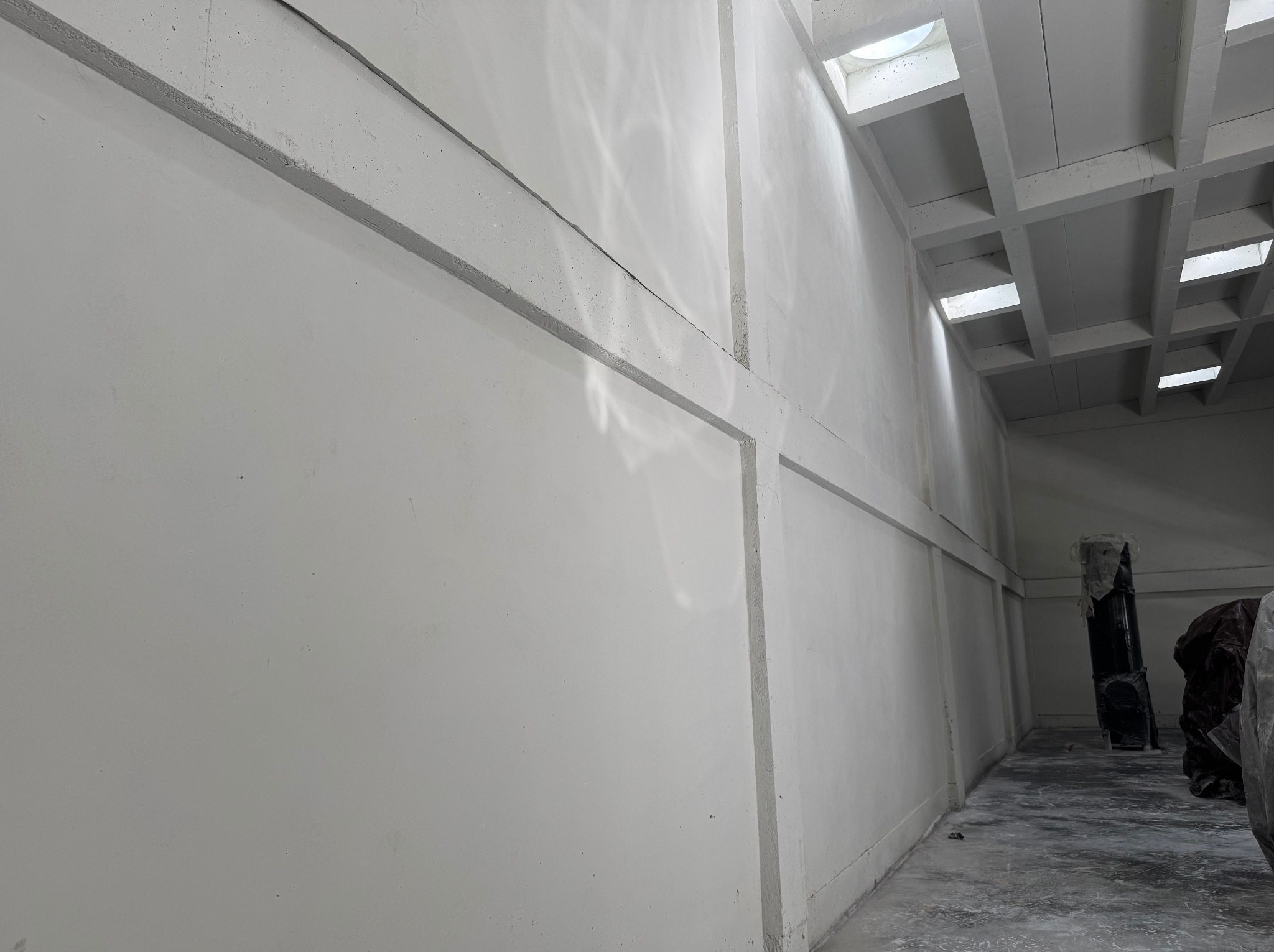 Wnętrze po tynkowaniu maszynowym: gładkie, białe ściany z widocznymi elementami konstrukcyjnymi i świetlikami w suficie. Sprzęt budowlany w tle.