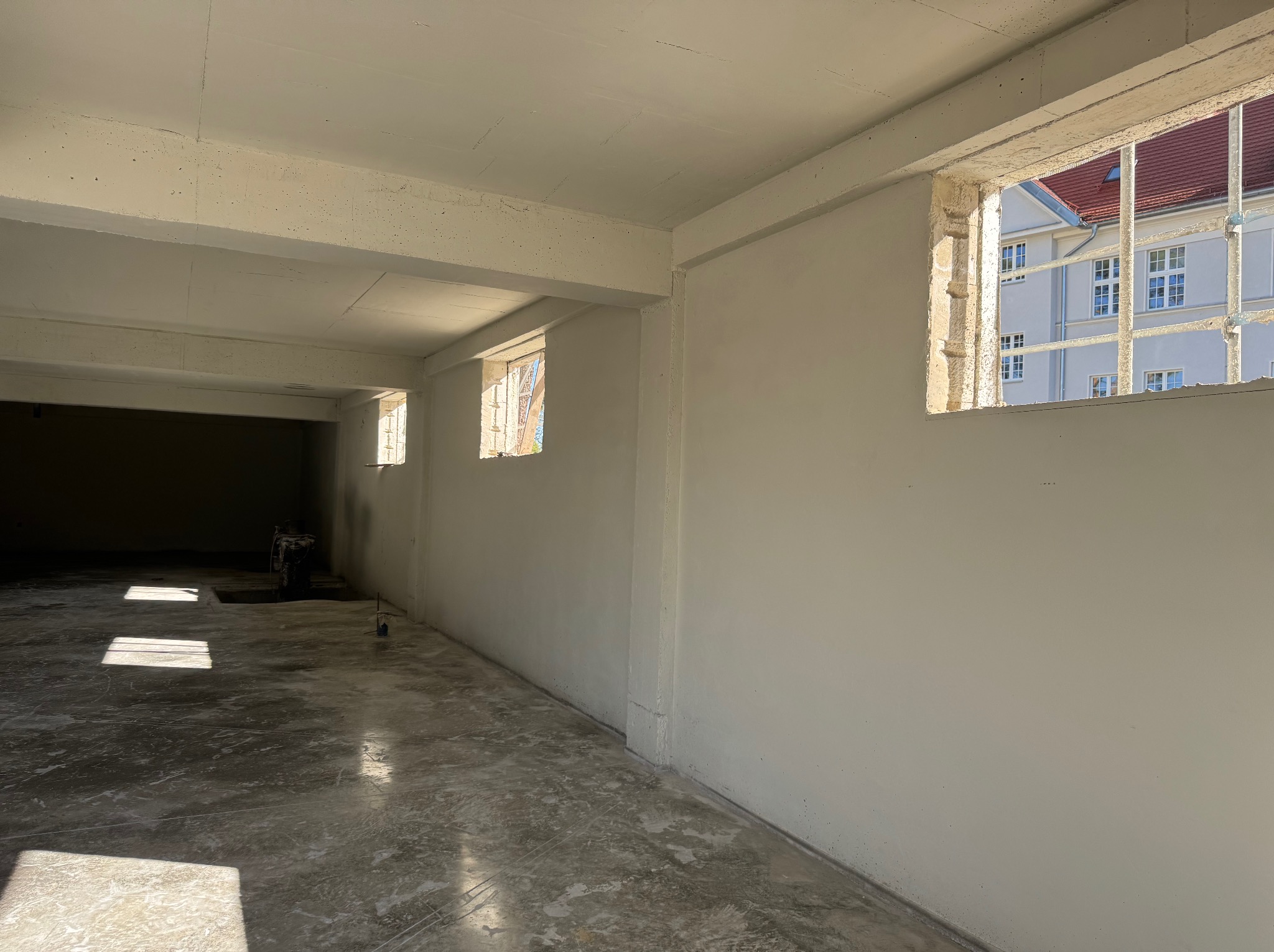 Wnętrze po tynkowaniu: jasne ściany, betonowa podłoga z refleksami światła słonecznego wpadającego przez okna z widokiem na budynek z czerwonym dachem.