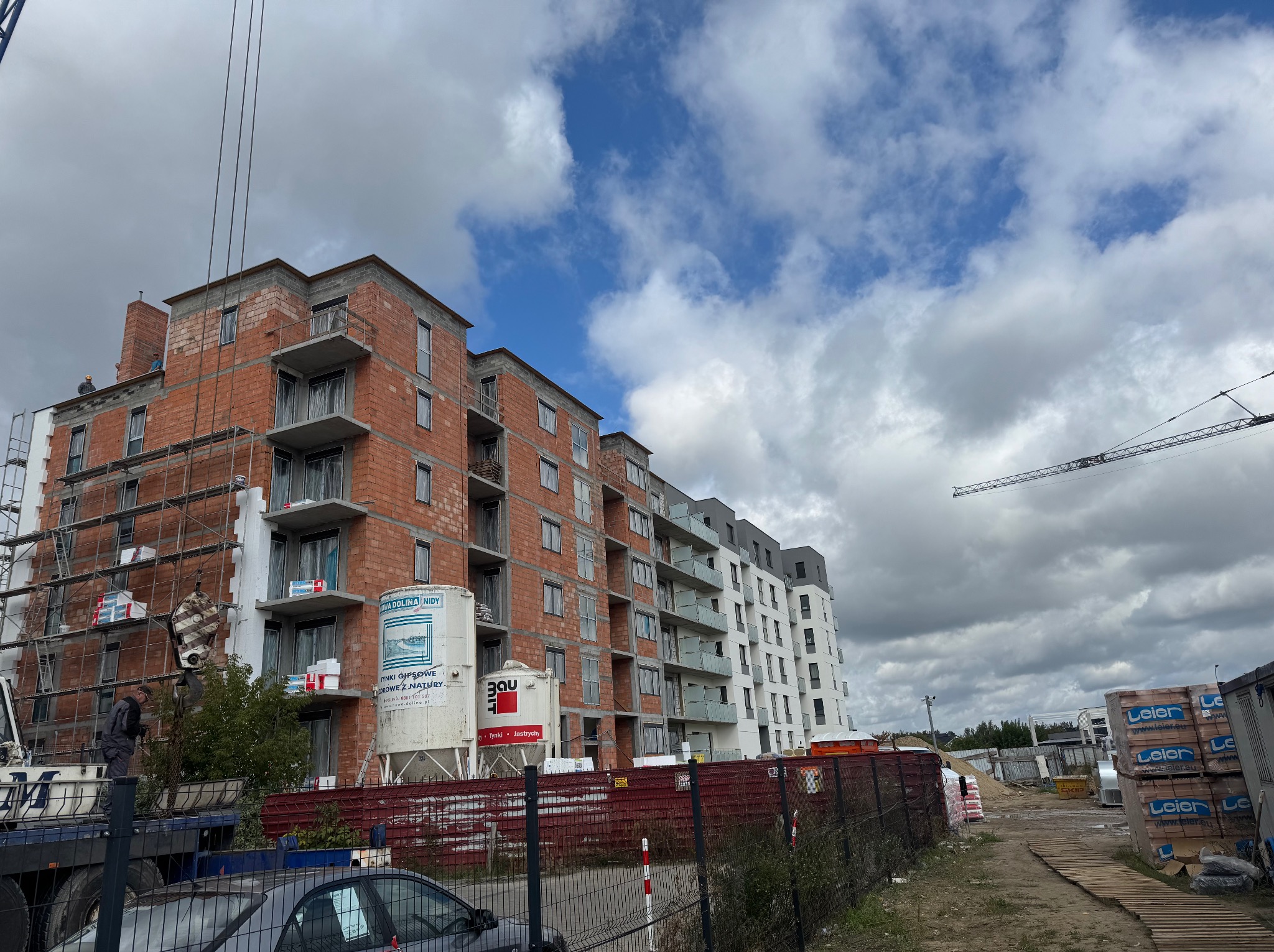 Budowa bloku mieszkalnego z cegły, widoczne rusztowania, zbiorniki na tynk i dźwig. Częściowo ocieplony budynek z balkonami na tle błękitnego nieba.
