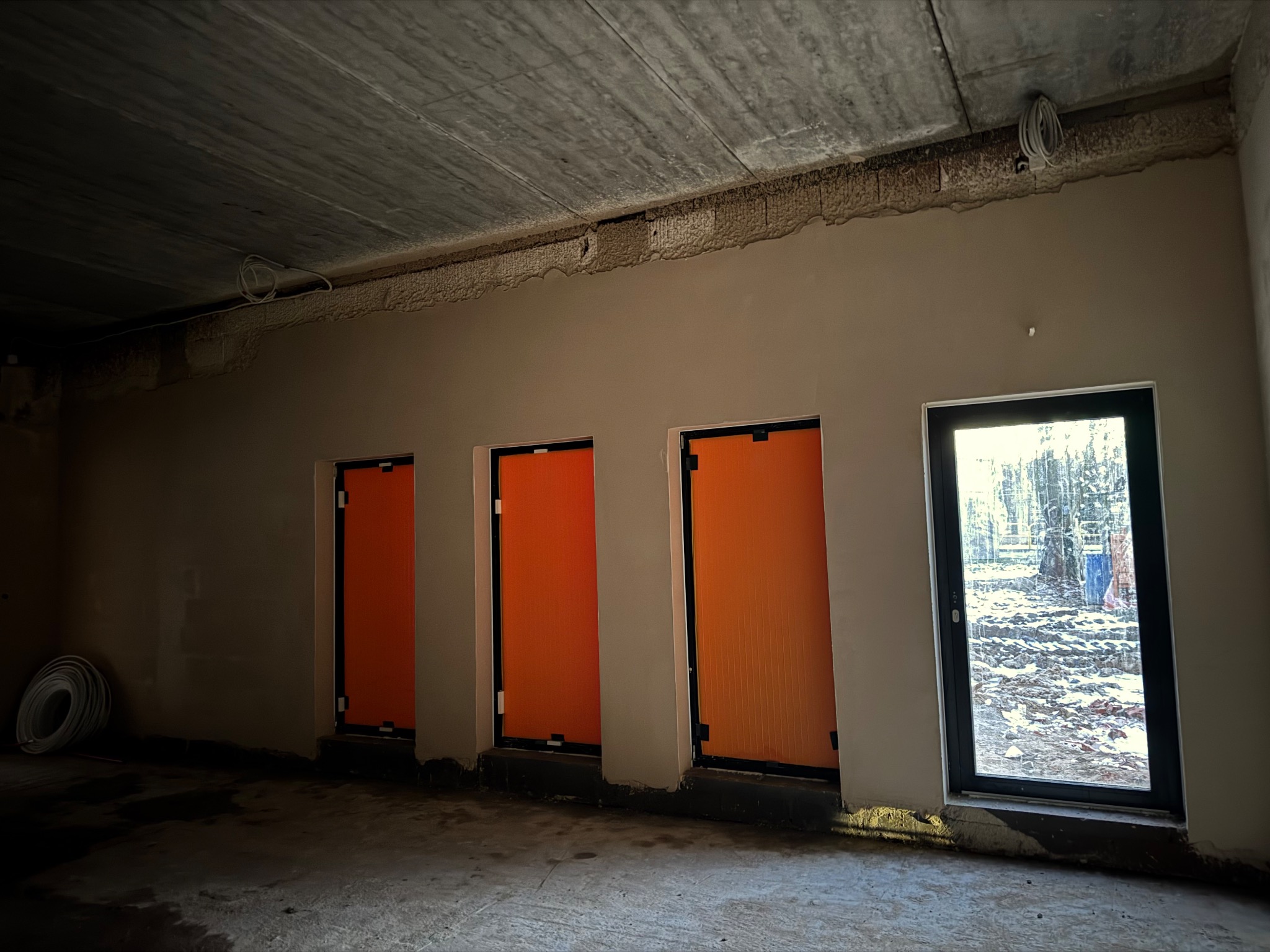 Surowy stan wnętrza z trzema pomarańczowymi drzwiami i jednym oknem z widokiem na zewnątrz. Widoczne niedoskonałości tynku i betonowy sufit.