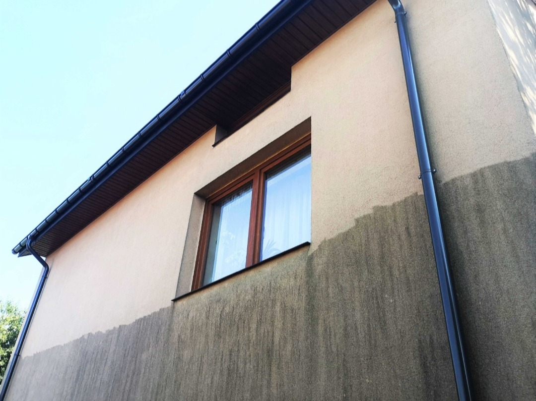 Fragment elewacji budynku z widocznym efektem czyszczenia, dolna część ściany ciemniejsza od górnej, okno z drewnianą ramą i rynna wzdłuż rogu budynku.