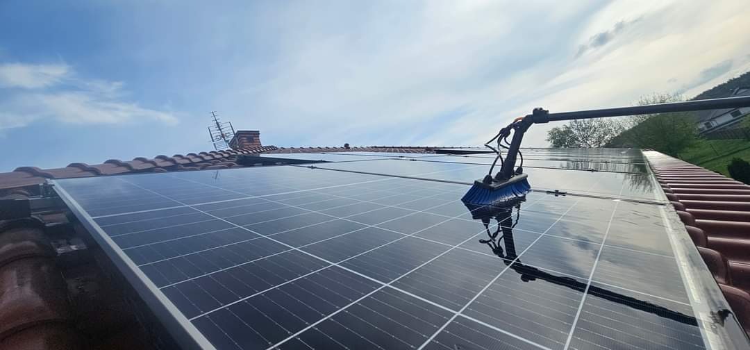 Czyszczenie paneli słonecznych na dachu domu za pomocą szczotki na teleskopowym wysięgniku; widoczna mokra powierzchnia paneli odbijająca niebo.