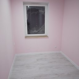 Marcin Wiewióra - Pomalowane na różowo wnętrze pokoju z białymi listwami przypodłogowymi i szarą podłogą, okno zabezpieczone folią.