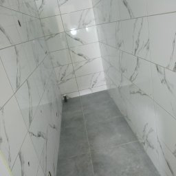 Marcin Wiewióra - Wnętrze łazienki po remoncie z wyłożonymi białymi płytkami imitującymi marmur na ścianach i szarymi płytkami na podłodze, widoczne otwory na armaturę.
