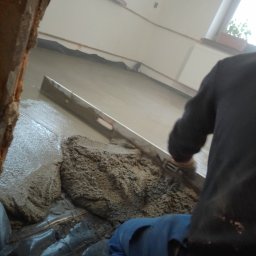 Marcin Wiewióra - Wylewka betonowa rozprowadzana łatą poziomującą na podłodze z siatką zbrojeniową, widoczny fragment ściany z cegły i okno z kwiatami.