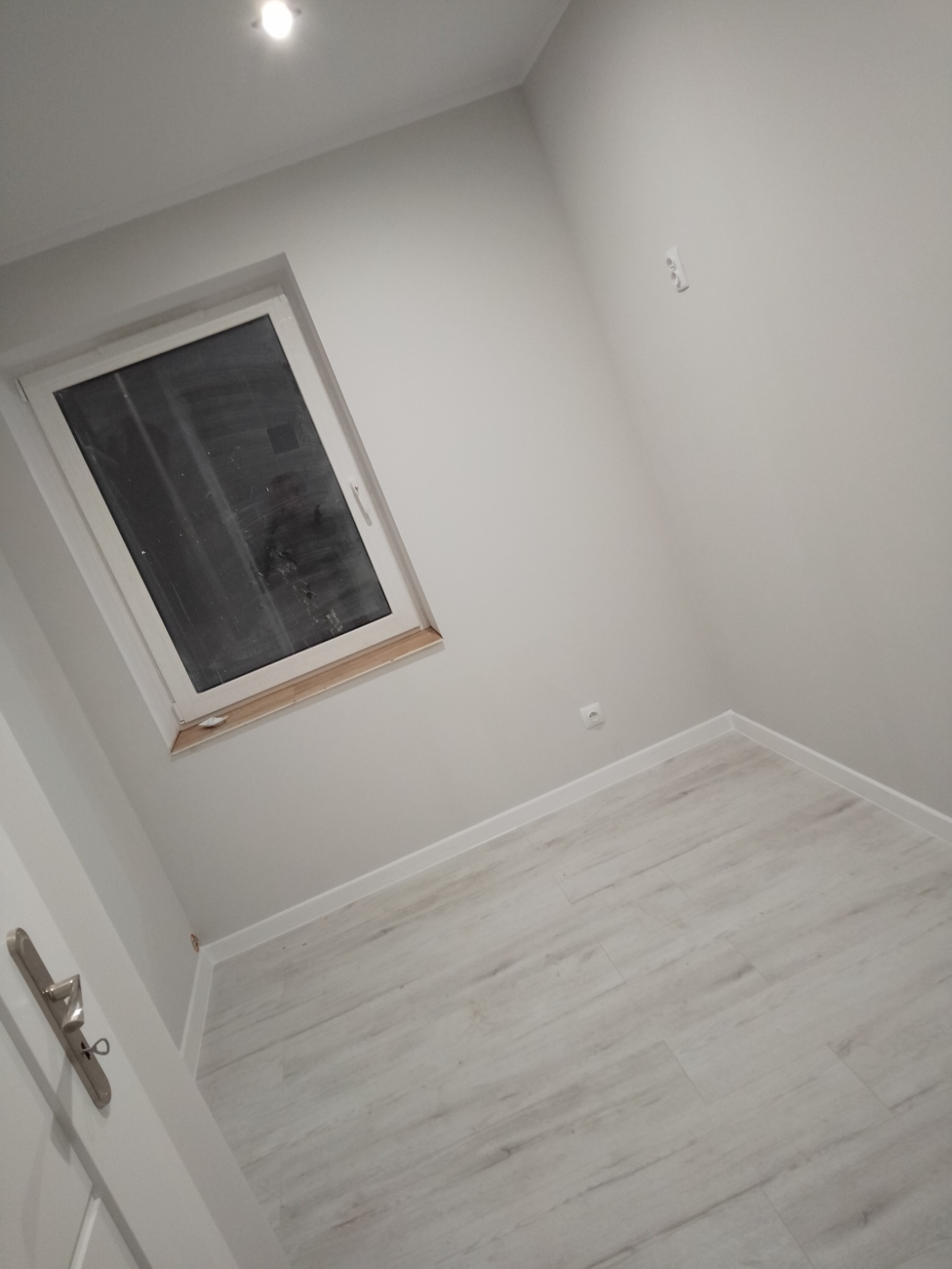 Jasne, świeżo pomalowane wnętrze pokoju z oknem dachowym, nową podłogą i listwami przypodłogowymi, widoczny fragment drzwi z klamką.