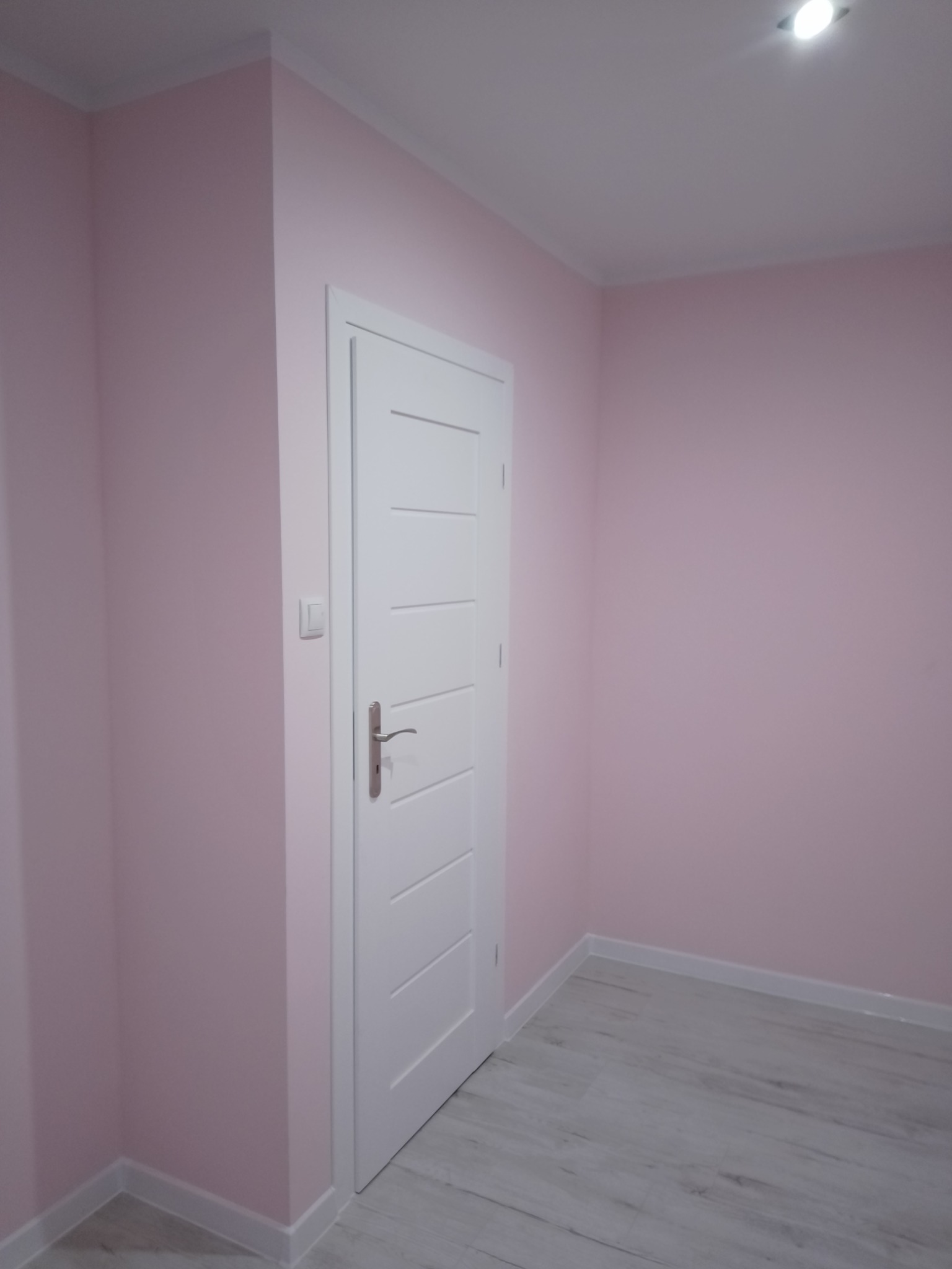 Pomalowane na różowo wnętrze pokoju z białą, nowoczesną stolarką drzwiową i listwami przypodłogowymi oraz jasną podłogą laminowaną imitującą drewno.