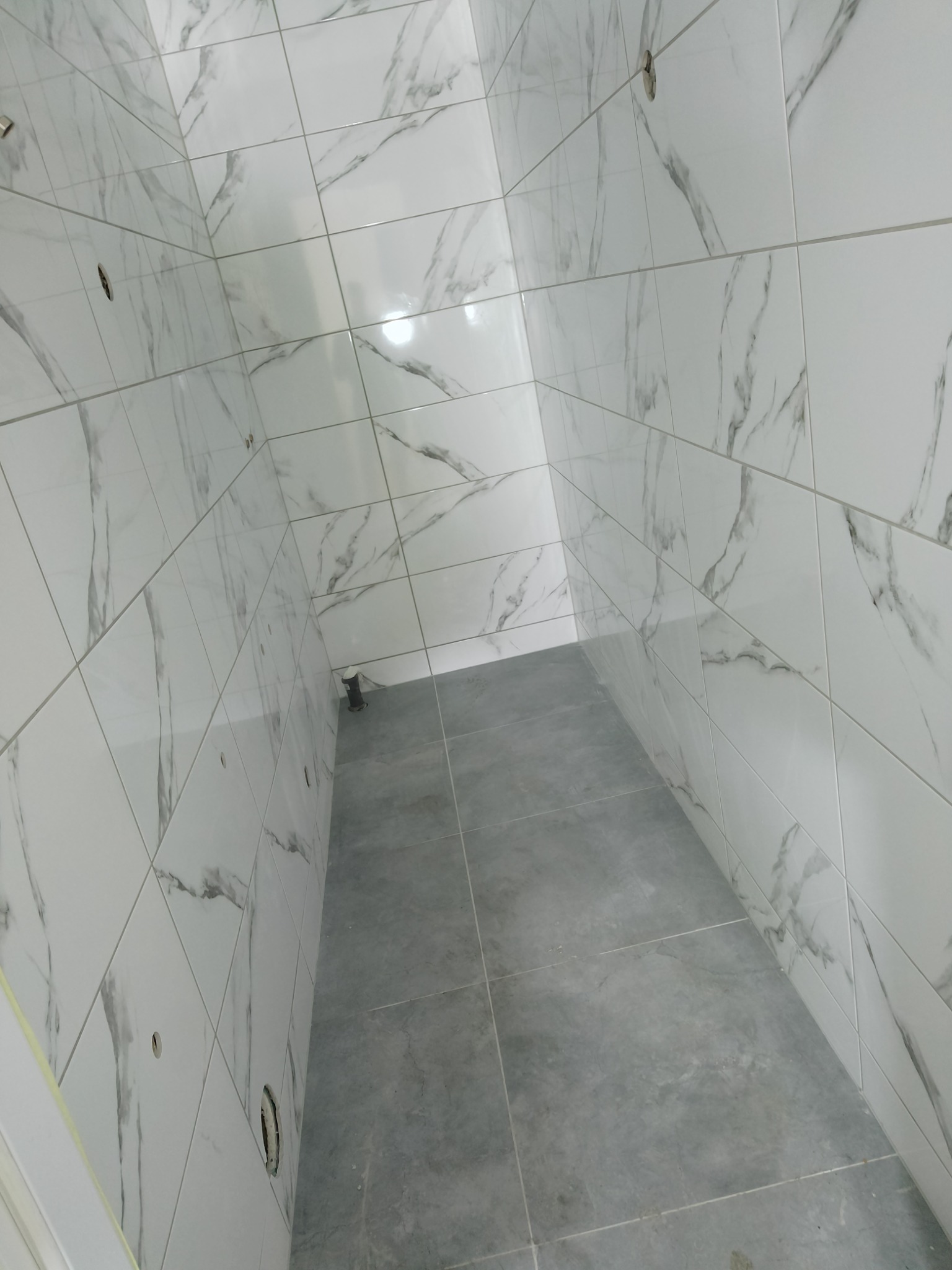 Wnętrze łazienki po remoncie z wyłożonymi białymi płytkami imitującymi marmur na ścianach i szarymi płytkami na podłodze, widoczne otwory na armaturę.