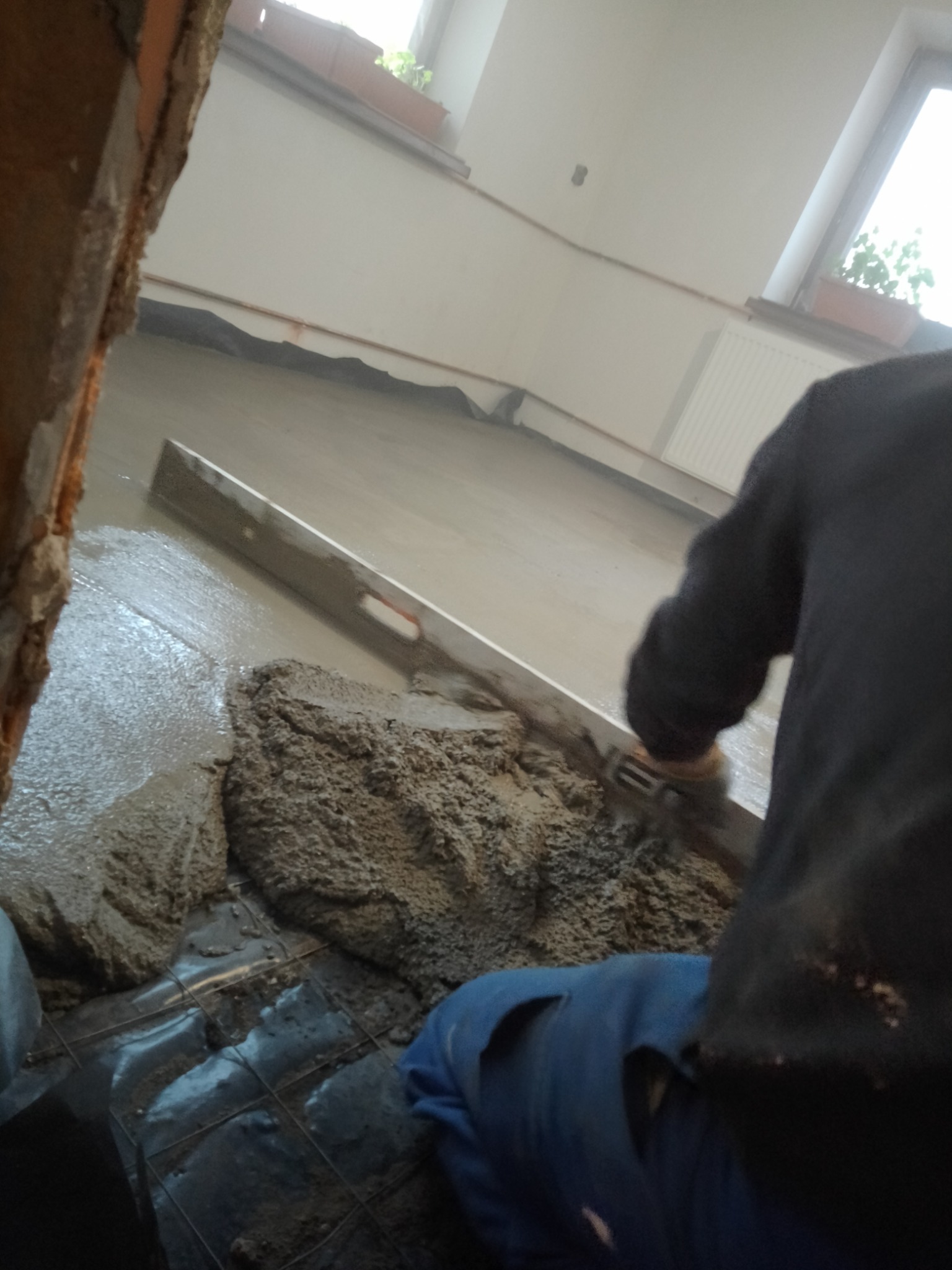 Wylewka betonowa rozprowadzana łatą poziomującą na podłodze z siatką zbrojeniową, widoczny fragment ściany z cegły i okno z kwiatami.