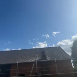 Ceglany budynek z nowym, brązowym dachem z blachy trapezowej w trakcie montażu, widoczne rusztowanie. Częściowo błękitne niebo z chmurami w tle.