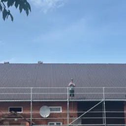 Nowy, brązowy dach z blachy falistej na ceglanym budynku. Na dachu stoi dekarz na rusztowaniu. Widoczne liście drzewa i błękitne niebo.