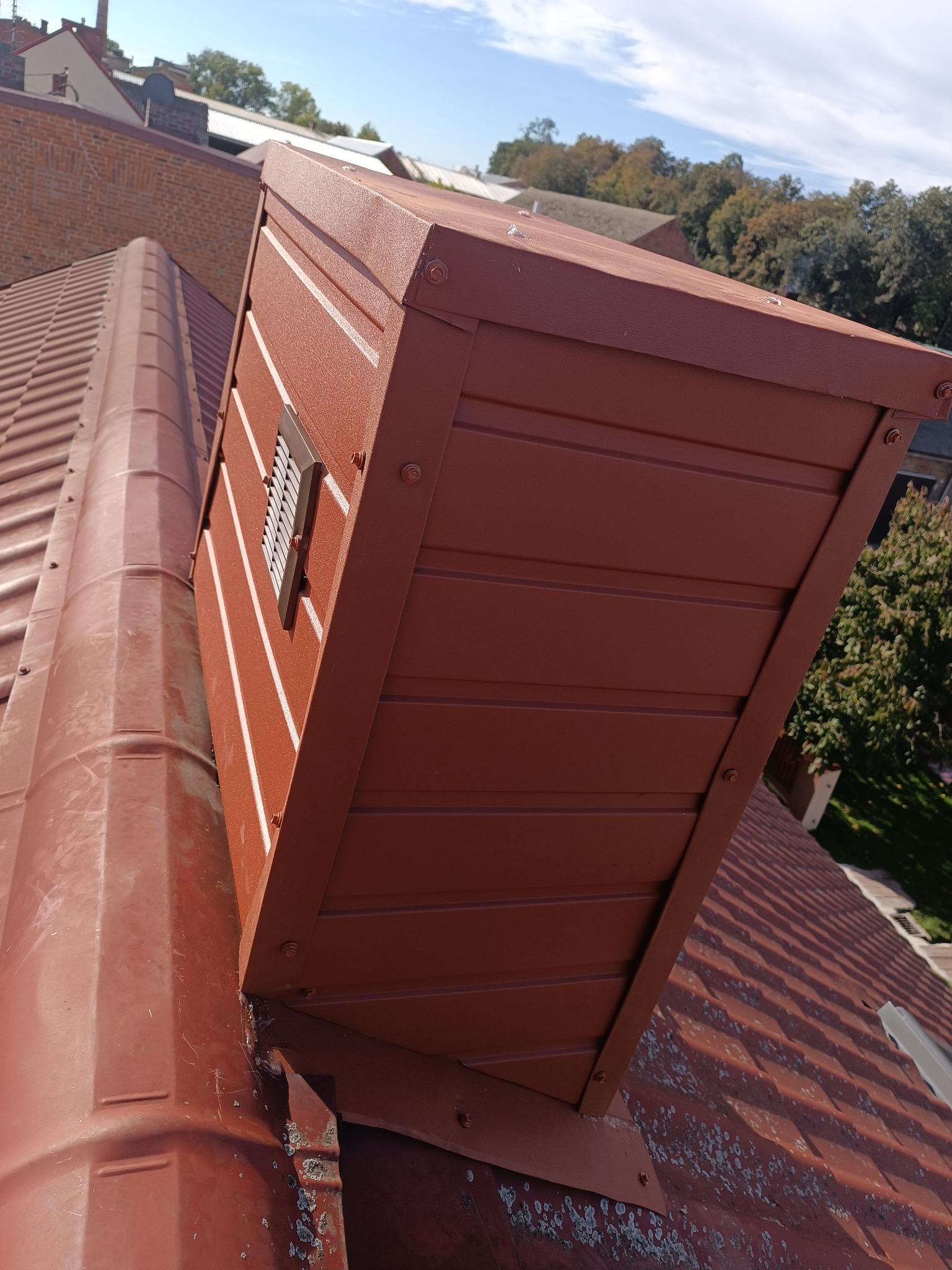 Brązowy komin wentylacyjny z kratką, zamontowany na dachu z blachy trapezowej w odcieniach brązu, na tle błękitnego nieba i zieleni drzew.