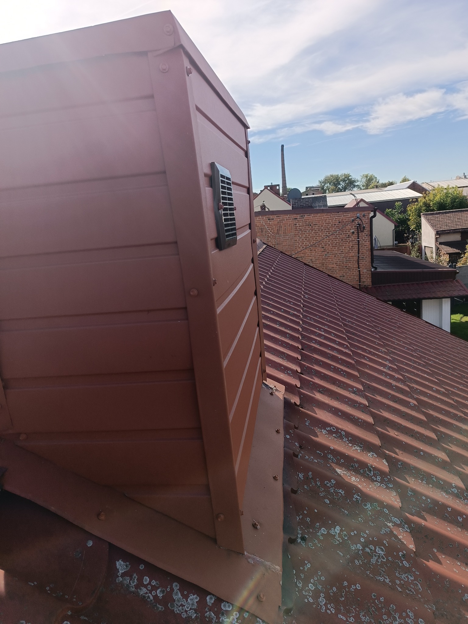 Nowy, brązowy dach z blachy trapezowej, z widocznym kominem wentylacyjnym i elementami mocującymi, na tle zabudowań i błękitnego nieba.