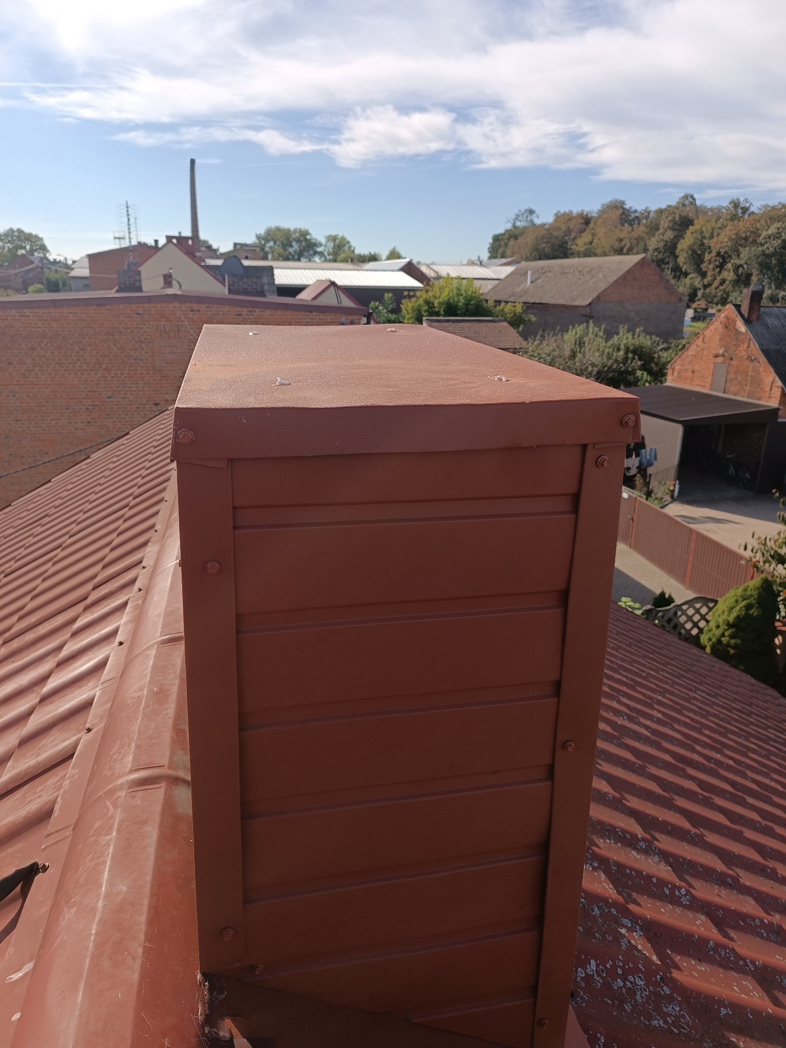 Czerwono-brązowy komin wentylacyjny z blachy na dachu z blachy trapezowej, widok z góry, w tle zabudowa mieszkalna i błękitne niebo.