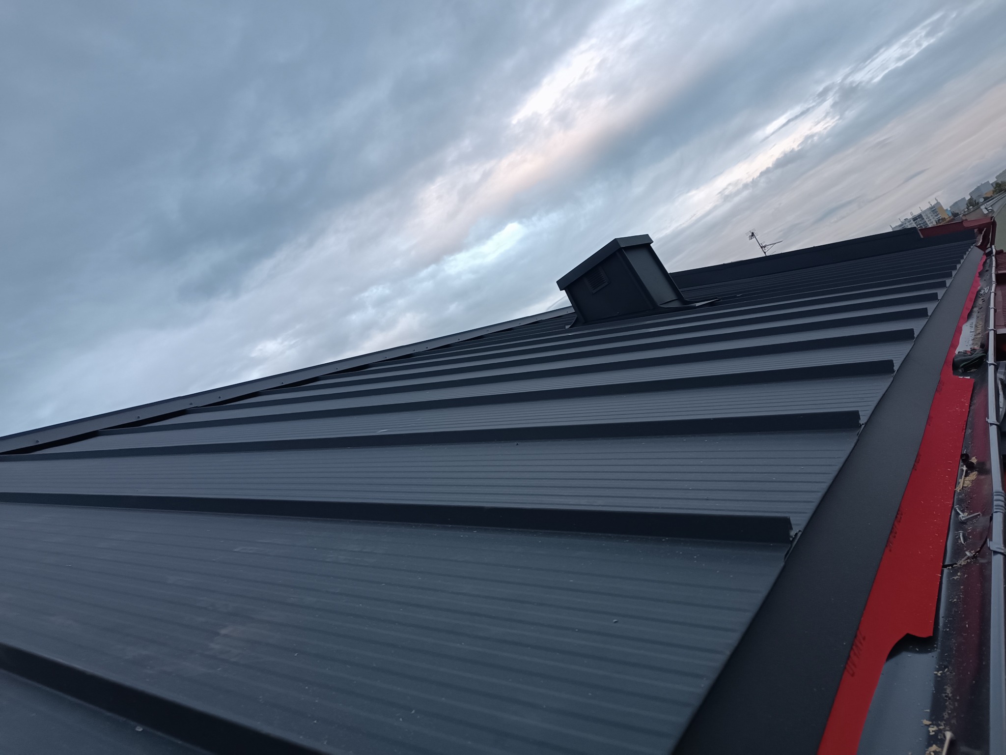 Nowy, grafitowy dach z blachy trapezowej, widziany pod kątem. W tle pochmurne niebo i fragment zabudowy miejskiej. Widoczny komin wentylacyjny i antena.